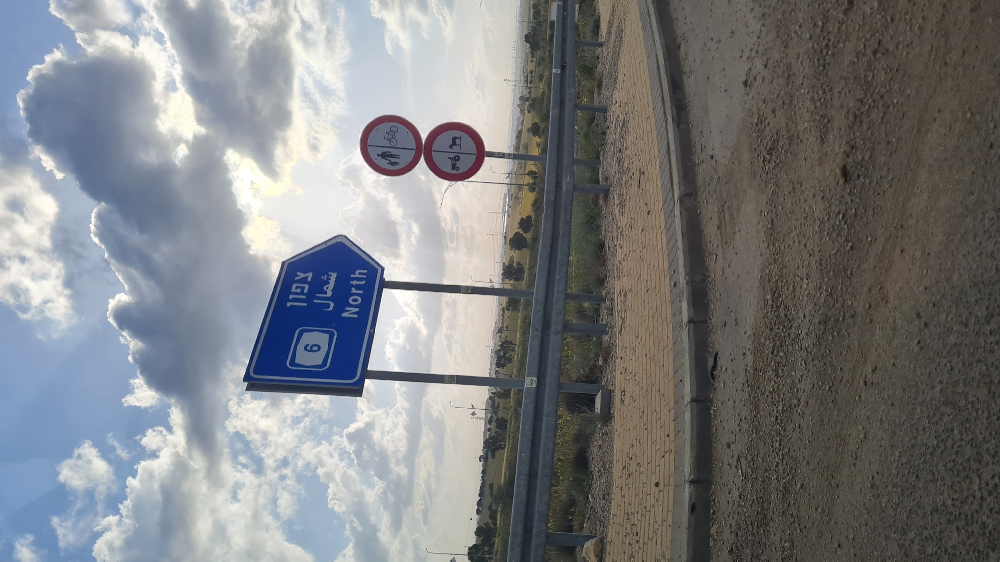 The image depicts a roadside scene in what appears to be the Bnei Shimon Regional Council, Israel. Road signs indicating Route 6 North are visible, along with signs prohibiting pedestrians, cyclists, and tractor-like vehicles. The environment consists of a roadside with a barrier, an adjacent field area, and a sky with scattered clouds. There are no people visible in the image.