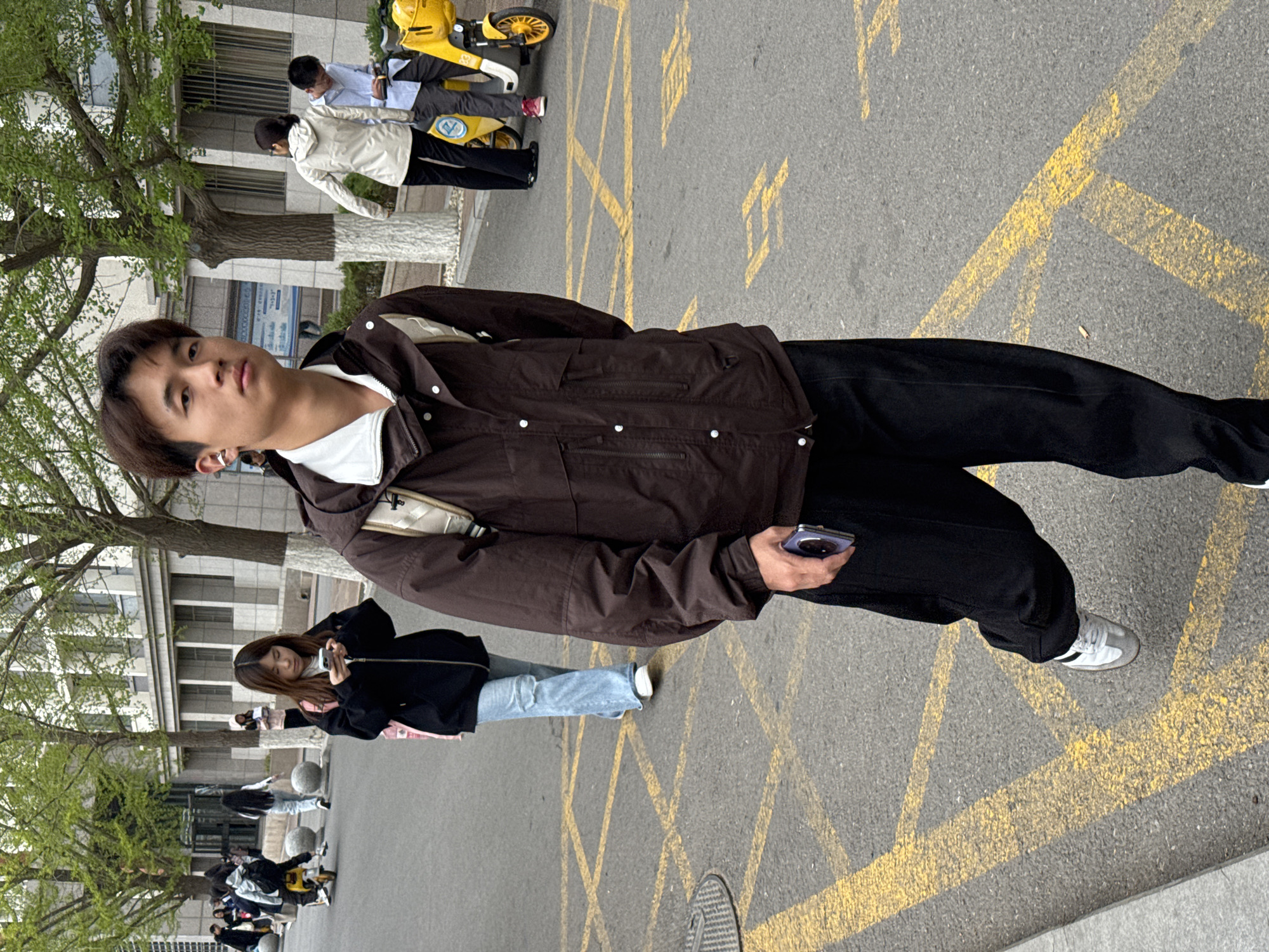 This image captures an outdoor scene in Zhongtun, China, featuring several people and urban elements. The primary subject is a young East Asian male, positioned prominently in the foreground. He is wearing a dark brown jacket over a white top, black pants, and white and grey sneakers. He has dark hair, an earring in his left ear, and holds a dark-colored smartphone in his right hand. He looks directly towards the camera with a neutral expression.
Behind him, to the left, another young woman with long dark hair is visible, wearing a dark jacket and light blue jeans, looking down at her own smartphone. Further in the background, several other individuals are present. Two people are near bright yellow shared bicycles; one in a light blue shirt appears to be sitting on a bike, while another in a white jacket stands beside it. More indistinct figures can be seen walking or standing further down the street.
The setting appears to be a paved street or a pedestrian area, possibly within a university campus or urban center. The ground is grey asphalt marked with distinctive yellow crosshatch patterns and other lines, typically indicating restricted zones. A round metal manhole cover is visible in the lower right corner. Lush green trees, some with white-painted trunks, line the left side of the scene, and multi-story buildings with windows are visible in the background. The lighting suggests it is daytime, possibly under overcast skies, given the soft shadows.
While specific brand names on the shared bicycles or detailed text on the buildings are not clearly legible, the yellow shared bikes are commonly found in Chinese cities. The yellow markings on the road resemble standard Chinese road signage. The overall atmosphere is casual and appears to depict daily life in an urban environment.