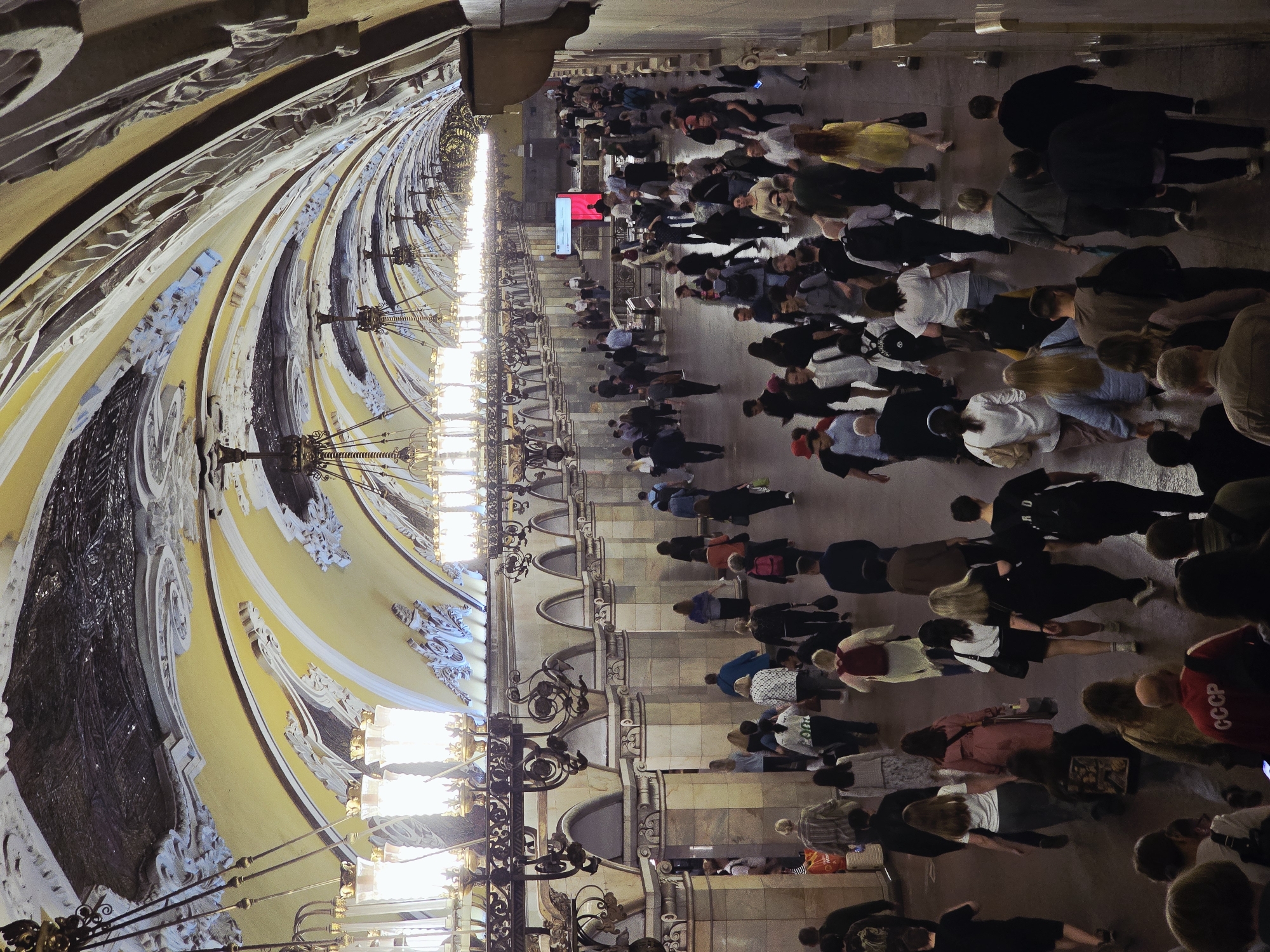 The image depicts a bustling subway station in Moscow, Russia. The architecture is ornate with curved ceilings decorated in shades of yellow, white, and dark gray. Intricate moldings and elaborate chandeliers adorn the space. A large crowd of people is moving through the station. Many are dressed in casual clothing. A red sign or screen is visible in the distance. The floor is paved with light-colored stone tiles.