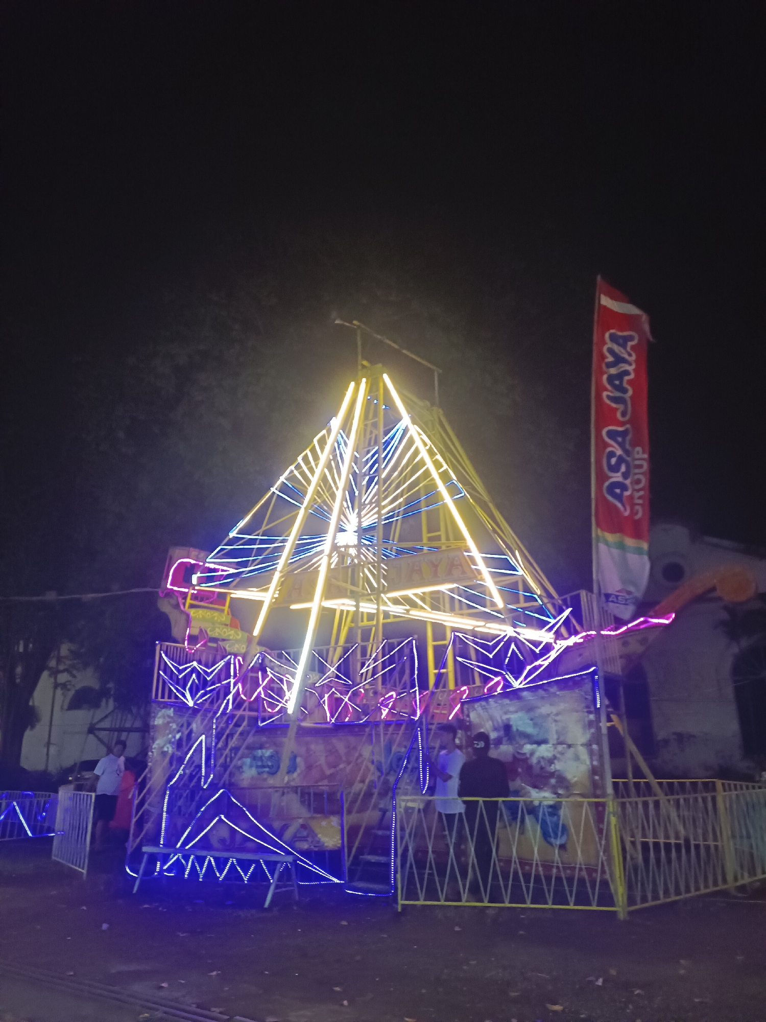 The image shows a small amusement ride, possibly a swing ride, brightly lit with yellow, pink, and blue lights. The ride has a central triangular structure with lights outlining its shape. A banner with "ASA JAYA GROUP" in red text on a white background stands to the right of the ride. The ride is surrounded by yellow fencing. Several people are standing near the ride. The scene appears to be at night in Tulangan, Indonesia.