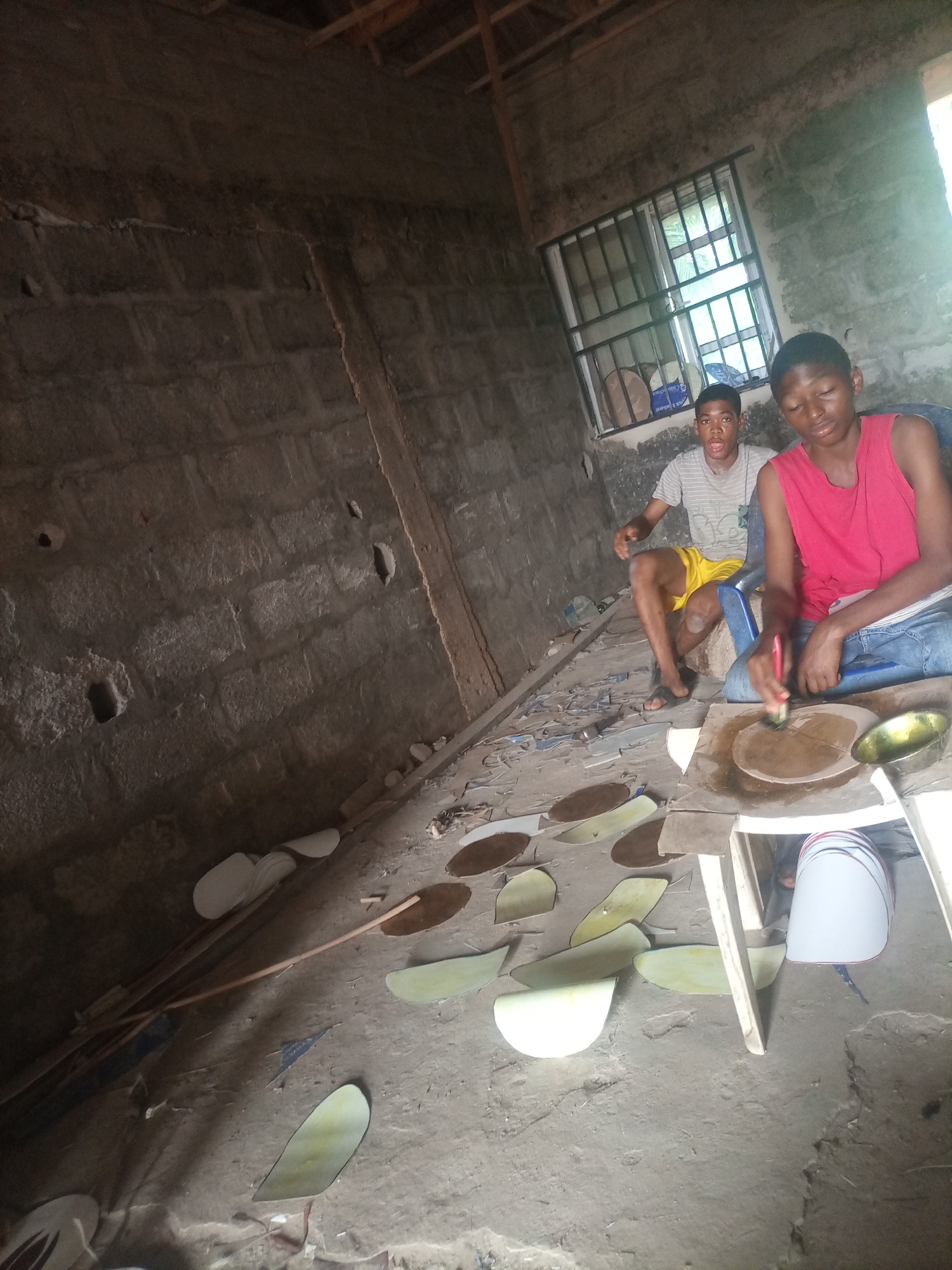 Two young men are working in an unfinished building. The subject on the right, wearing a red tank top and blue jeans, is focused on applying something to a flat, circular object with a brush. The object appears to be a piece of metal or wood that has been divided into sections. Next to it is a small, metallic bowl. The second subject, wearing a striped t-shirt and yellow shorts, sits a short distance away, looking towards the viewer with a surprised or excited expression.
The setting is a rudimentary interior space with bare concrete block walls, a dirt floor, and exposed wooden rafters in the ceiling. A window with metal bars is visible on the back wall. Scattered on the floor are numerous flat, leaf-shaped objects, some in shades of brown and others in a pale green or yellow. There are also long, thin wooden poles leaning against the wall.
The overall impression is of a workshop or construction site where a craft is being practiced. The lighting suggests it is daytime, with natural light coming from the window and possibly an open entrance not fully visible. The atmosphere seems industrious, with one individual actively engaged in the task and the other observing or pausing. The location context of Nkpor, Nigeria, places this scene in a specific geographic and cultural setting, though no explicit visual cues within the image directly confirm this beyond the general appearance of the construction and the subjects. No visible text is present in the image.