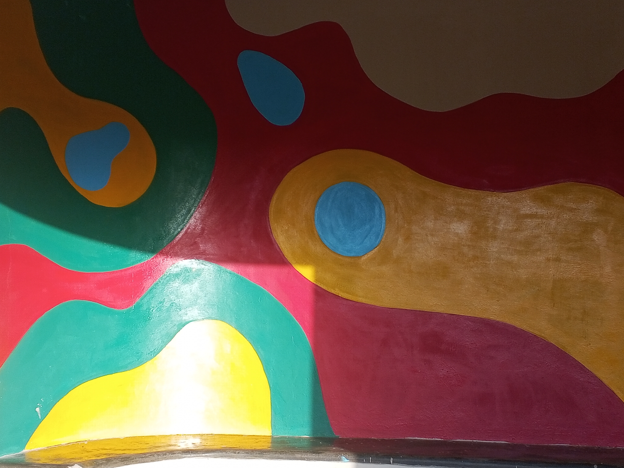 A mural painting is depicted on a wall. The painting features abstract shapes and patterns in several different colors, including red, green, yellow, orange, light brown, and blue. The colors are arranged in a swirling, organic manner. Light is visible shining on the bottom of the mural. The location is Tawpo, Nigeria.
