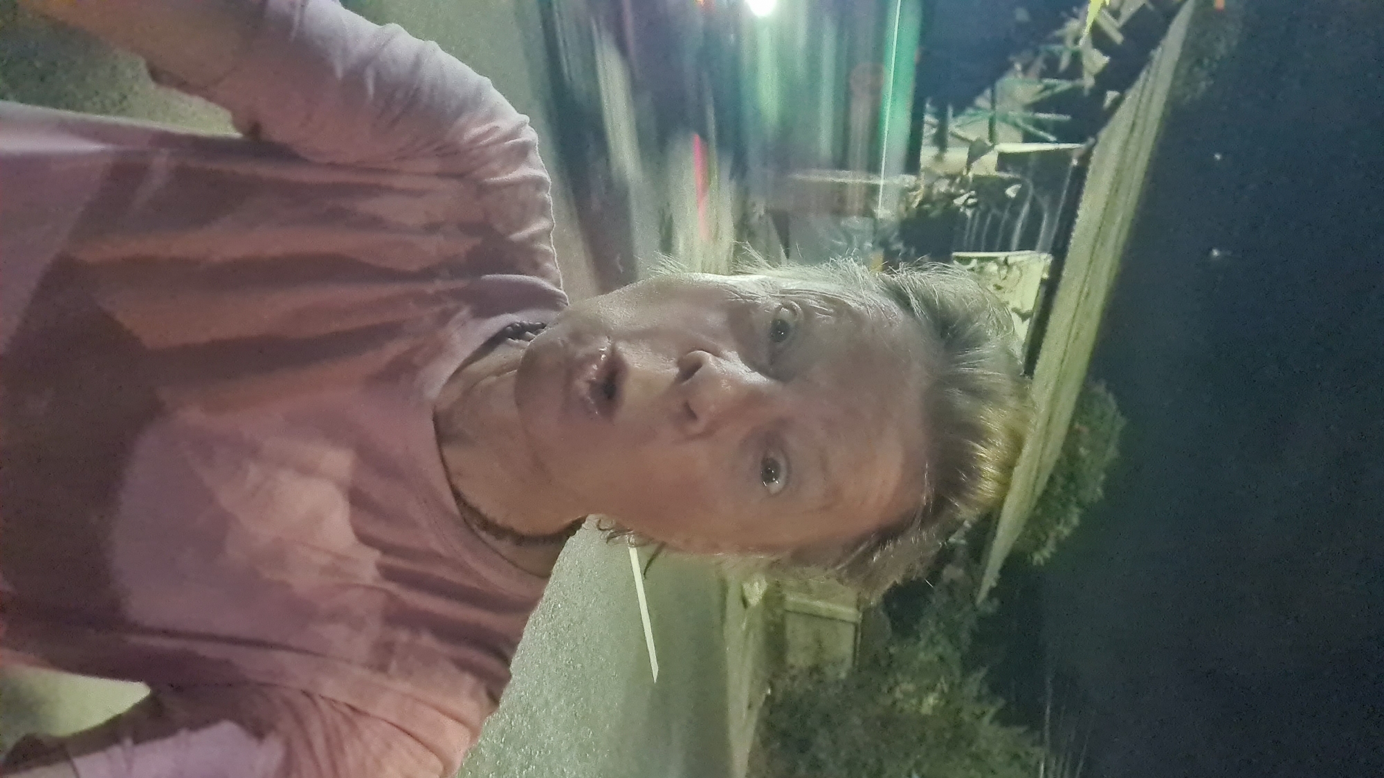 A woman with blonde hair pulled back stands in a selfie, making a surprised expression with her mouth open and eyes wide. She is wearing a pink t-shirt and a dark necklace. The background shows a street at night, with a blurry car on the left and a dilapidated building with a tin roof on the right. Trees and foliage are visible behind the building. A white line marking the edge of the road is visible in the foreground. The scene appears to be on a street in Denpasar, Indonesia, evidenced by the common architectural style of the building and the tropical vegetation.