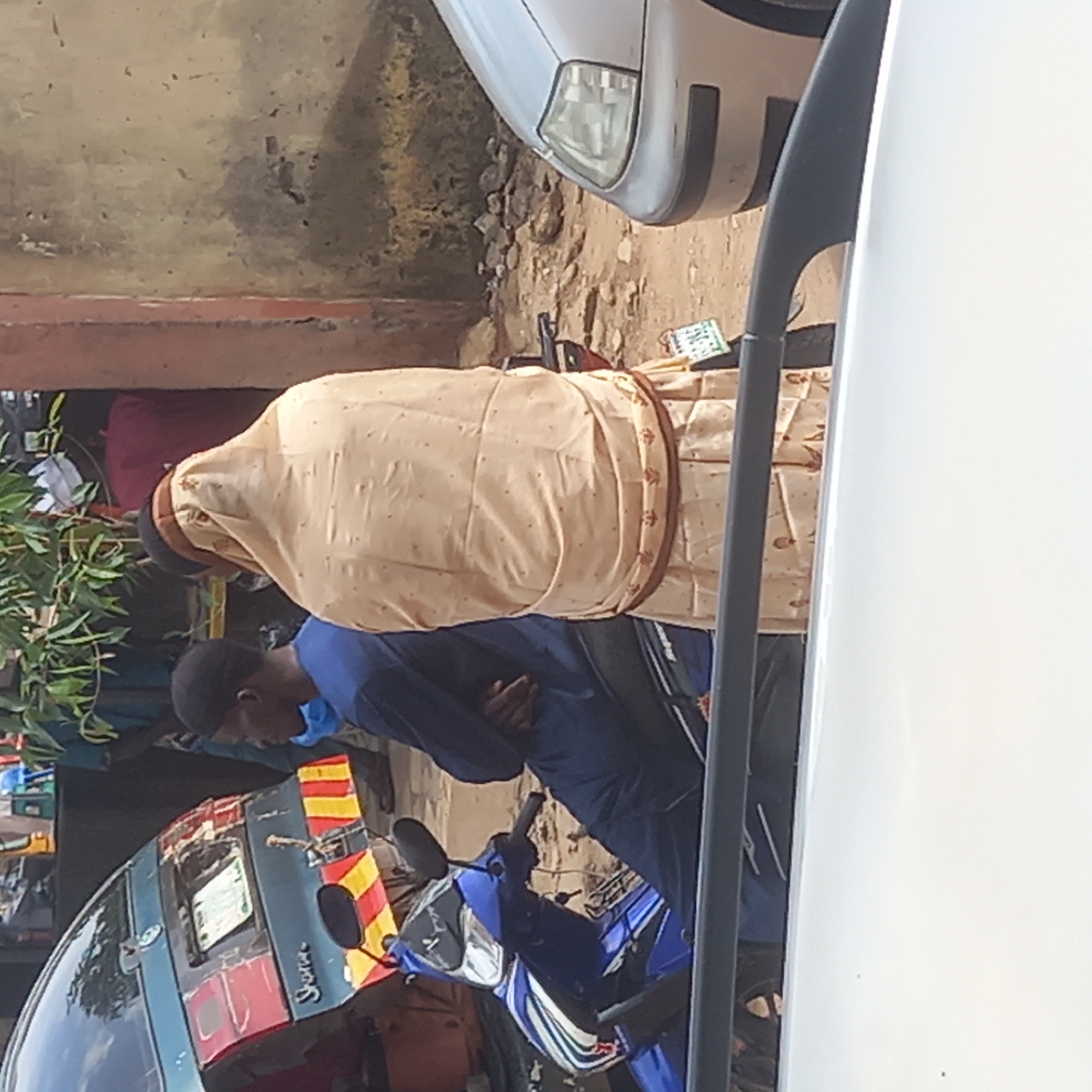 The image captures a street scene in what appears to be Sardauna, Nigeria. A man in a blue shirt and face mask stands near a parked blue and white motorcycle and a blue and red vehicle marked with yellow stripes. Adjacent to him is a person wearing a cream-colored garment with brown trim, possibly a religious robe or attire. To the right, partially obscured, is a white vehicle, its side prominently visible. In the background are elements of a building and greenery, adding to the context of an urban setting.