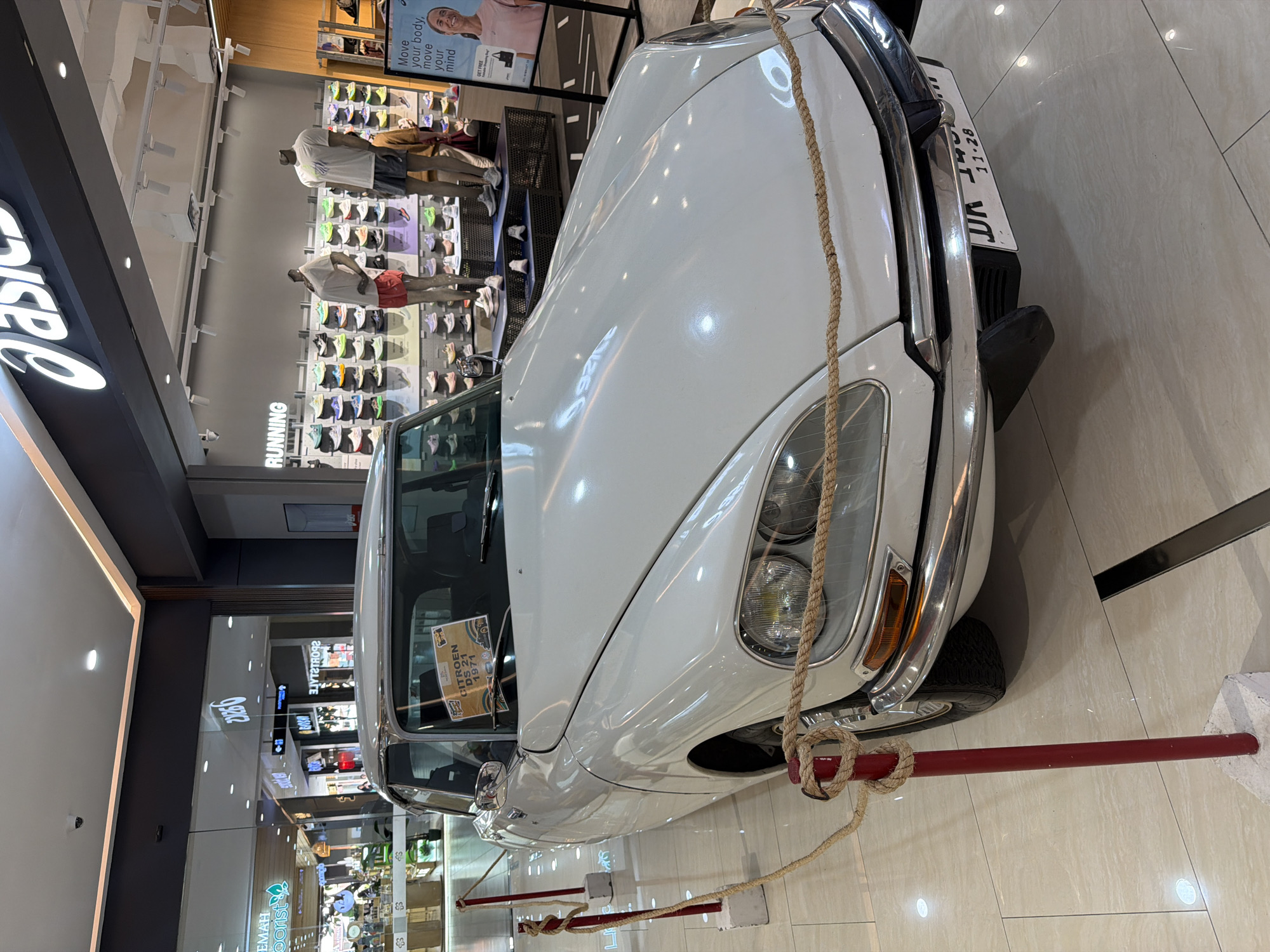 A white vintage Citroën DS 21, manufactured in 1971, is prominently displayed indoors, likely within a shopping mall in Kuta, Indonesia. The car is secured by ropes attached to red posts, preventing close access. A sign on its windshield clearly identifies it as a "CITROËN DS 21 1971." A partially visible license plate reads "UR 126" and "11-28."
The car is positioned on a tiled floor, reflecting the overhead lights of the mall. In the background, several retail stores are visible. To the left of the car, a large "ASICS" logo is partially seen, and further back, the word "RUNNING" is displayed on a wall. Mannequins wearing sportswear are visible inside the Asics store, surrounded by racks of athletic shoes. A poster within the store features a smiling woman and text that reads, "Move your body, move your mind" and "GET FREE."
Further into the mall, reflections show other store signs, including "EMAH florist" and "Yova" (reversed), indicating a diverse retail environment. The setting suggests a promotional event or exhibition showcasing the classic car within a modern shopping complex.