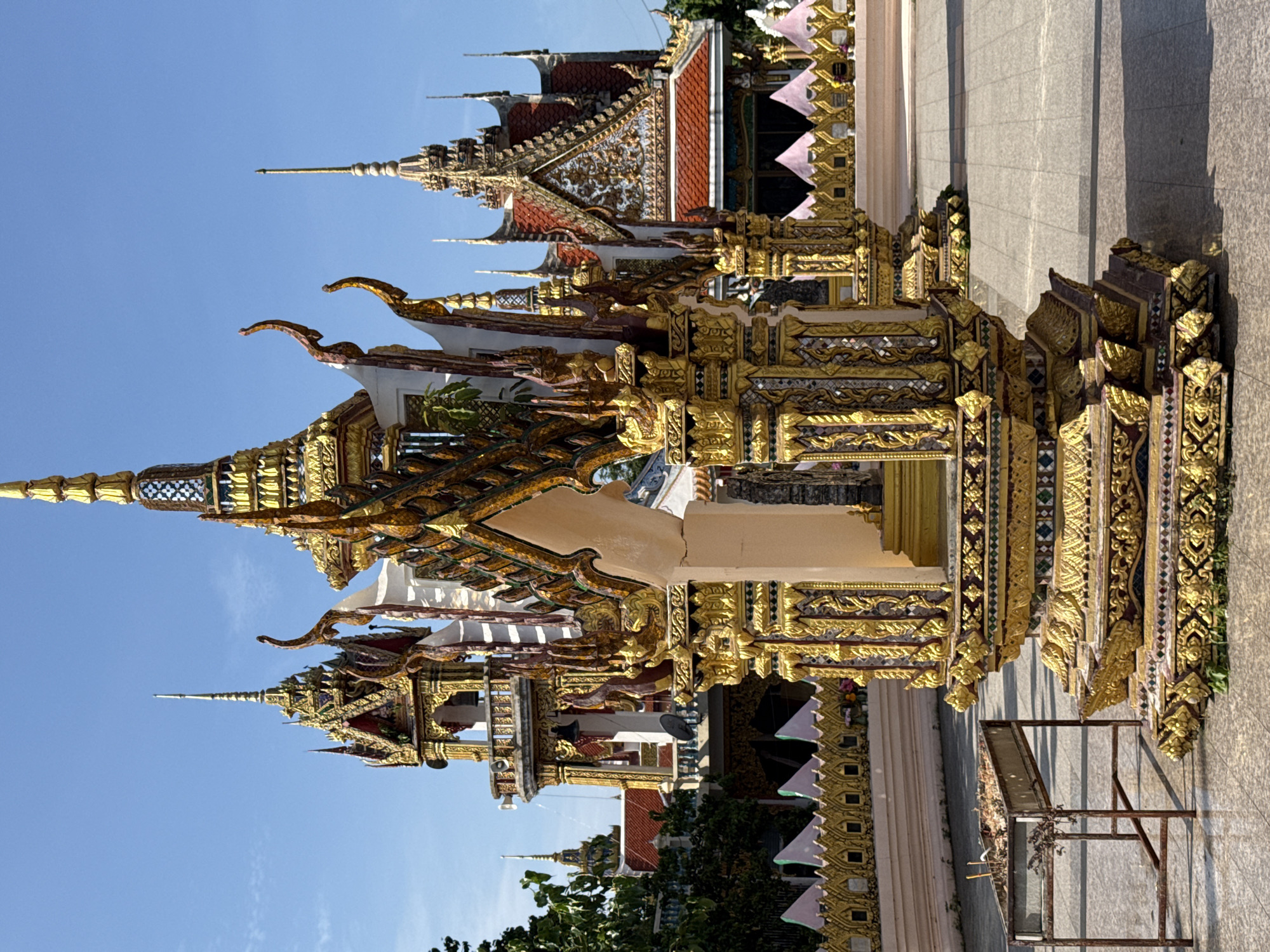 This is a daytime, medium shot photograph taken at Wat Phra Yai (Big Buddha Temple) in Ko Samui District, Thailand. The foreground features a highly ornate, gilded structure with multiple tiers and intricate decorative carvings, likely a shrine or altar. To the left of this structure, a simple, rusty metal brazier stands on the tiled ground, containing what appear to be burnt incense sticks and dried foliage. In the background, a prominent, multi-tiered stupa with a golden spire dominates the scene, flanked by other temple structures exhibiting similar ornate architecture and golden embellishments. The temple complex is situated outdoors with a clear blue sky overhead. The overall environment suggests a place of religious significance and cultural importance.