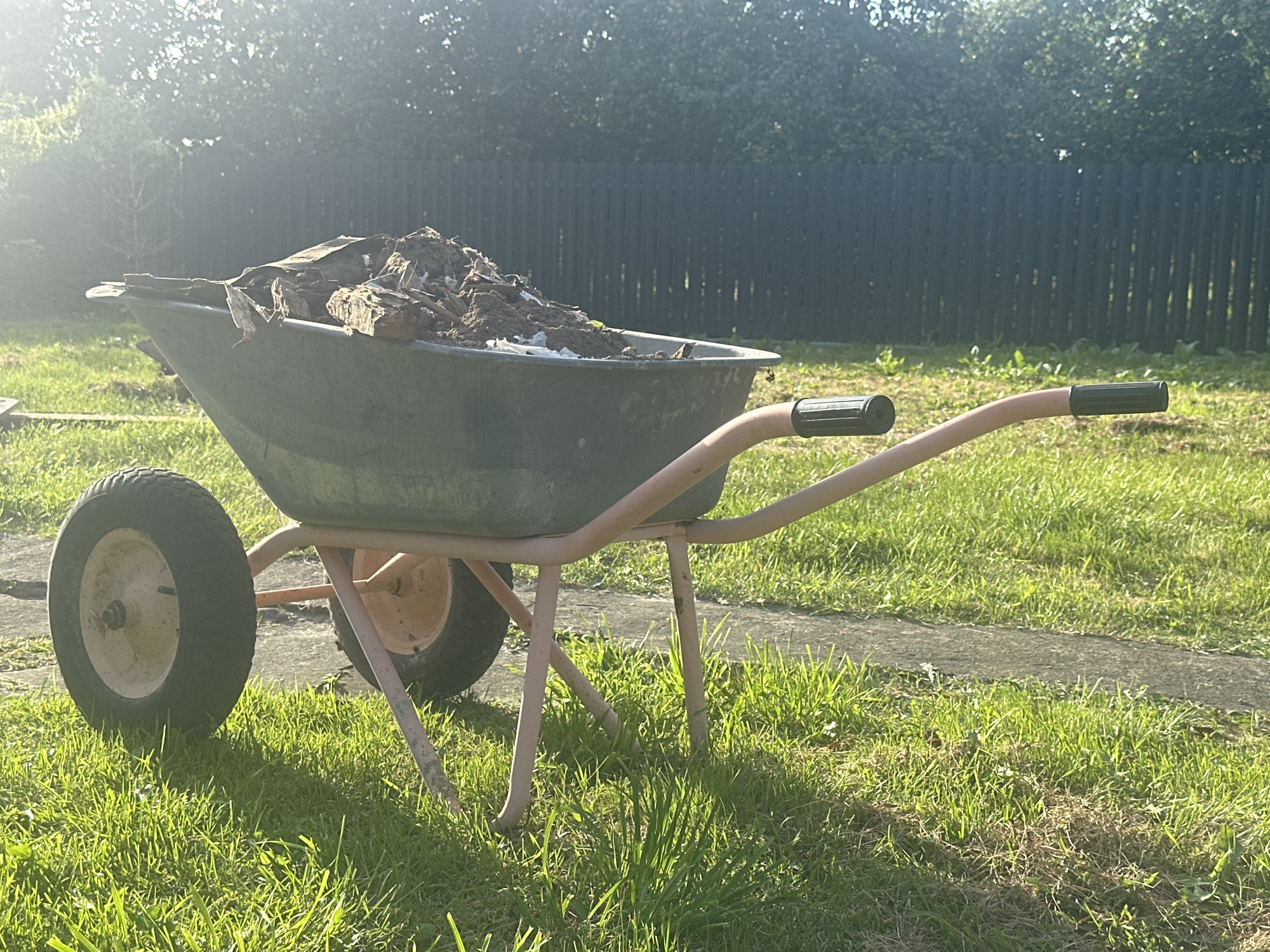 The image shows a green wheelbarrow filled with dirt and debris, including pieces of wood. The wheelbarrow has a light pink frame and two wheels, one larger than the other. It is sitting on a grassy lawn next to a paved path. In the background, there is a dark wooden fence and trees. The sun is shining, casting long shadows. The scene appears to be in a residential area, possibly a backyard, in the village of Mihanaŭicki sel’ski Savet, Belarus. There are no people visible in the image.