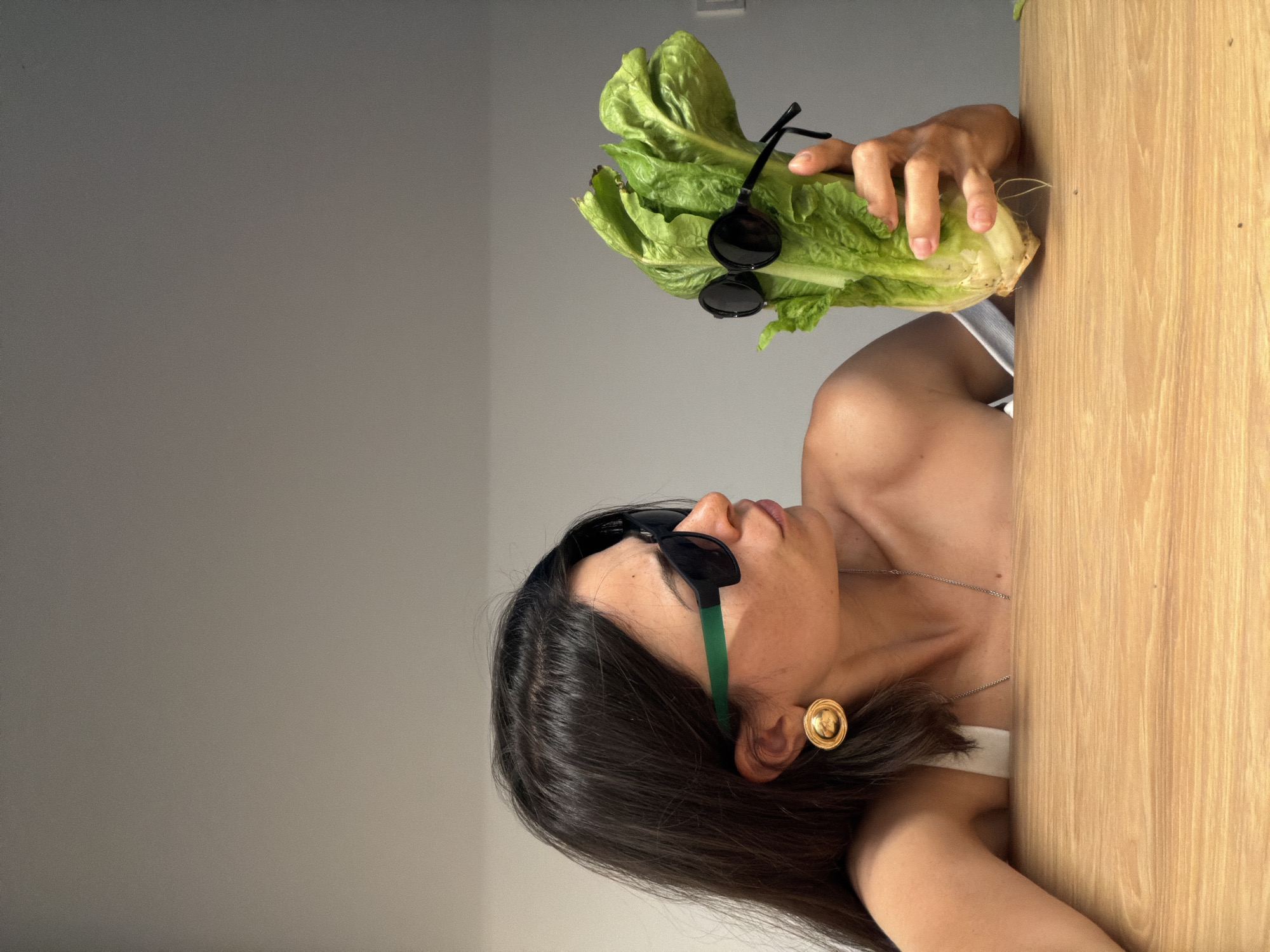 A medium shot of a woman holding a head of romaine lettuce with a pair of round black sunglasses hanging from it. The woman is wearing another pair of sunglasses with green frames and has an ornate gold earring. She also wears a thin necklace. The background consists of a light grey wall to the left and a light brown wooden surface to the right. The image is rotated counterclockwise. The location in the city of Budva, Montenegro cannot be determined.