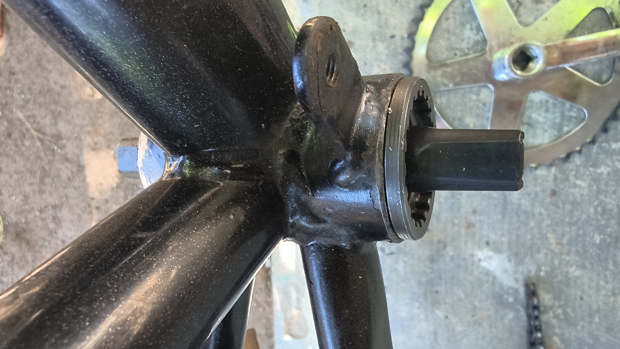 The image shows a close-up, low-angle shot of a bicycle's bottom bracket and crankset. The bottom bracket shell is black and appears to be welded to the bicycle frame, which is also black. A square spindle protrudes from the bottom bracket, and a silver crank arm with a chainring is visible to the right. The background consists of a grey concrete surface, with some green and yellow colors indicating foliage or a bicycle gear. The scene is outdoors and appears to be in an urban or suburban setting. There are no people visible in the image. There is no information in the image that indicates the location in the city of Mikhaylovka, Russia.