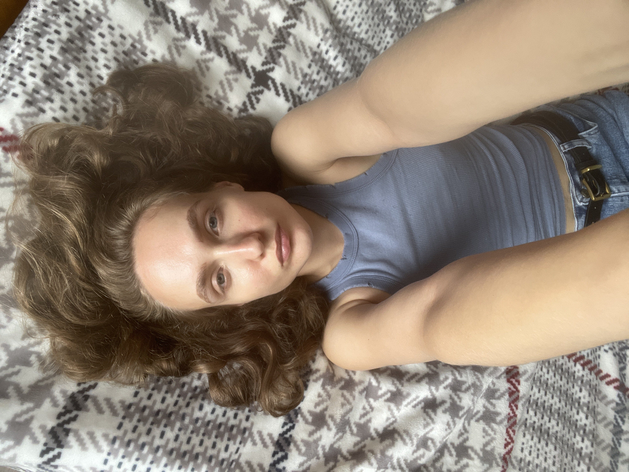 A young woman with long, wavy brown hair lies on her back on a patterned blanket. She is wearing a light blue ribbed tank top and blue denim shorts with a black belt. Her right arm is extended towards the camera, holding it. The blanket has a repeating geometric pattern of grey and white shapes. The image appears to be a selfie taken indoors, possibly in Sharkan, Russia, given the information provided, though the specific location is not visually discernible from the image itself.