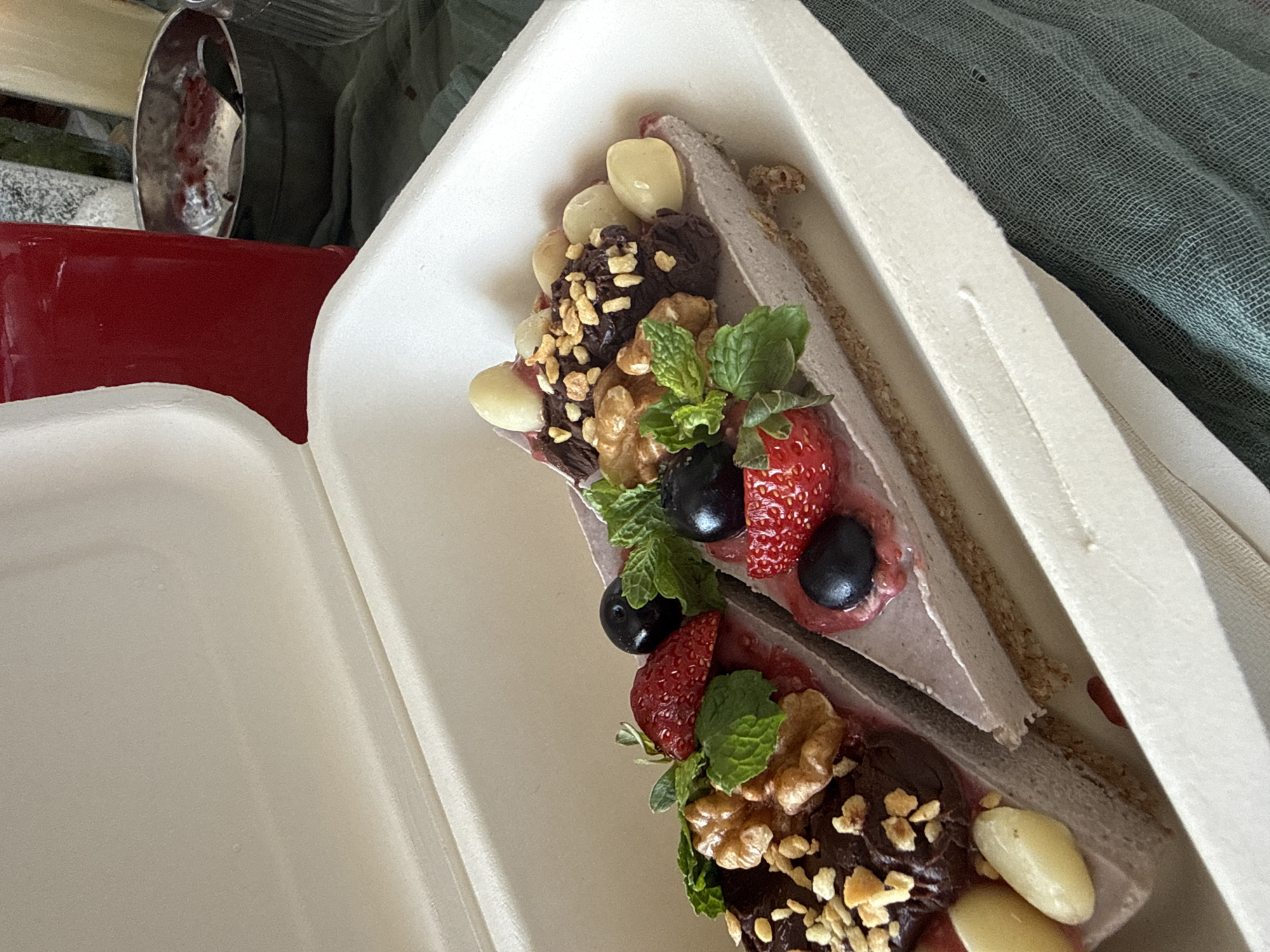 Two slices of raw vegan cheesecake sit side-by-side in a white takeout container. Each slice is topped with fresh berries, mint leaves, walnuts, and pale, oval-shaped nuts, possibly macadamia nuts or lychee nuts. The cheesecake itself appears to have a light purple hue, suggesting a berry or ube flavor, and a crumbly crust. The container is open and positioned on a dark green textured fabric, possibly a tablecloth or napkin. In the background, out of focus, is a tall red glass filled with a beverage and a metal bowl containing a dark red substance and a spoon. The overall scene suggests a dessert being served or transported. The image does not provide any visual cues to determine its specific location within Bac Nha Trang Ward, Vietnam.