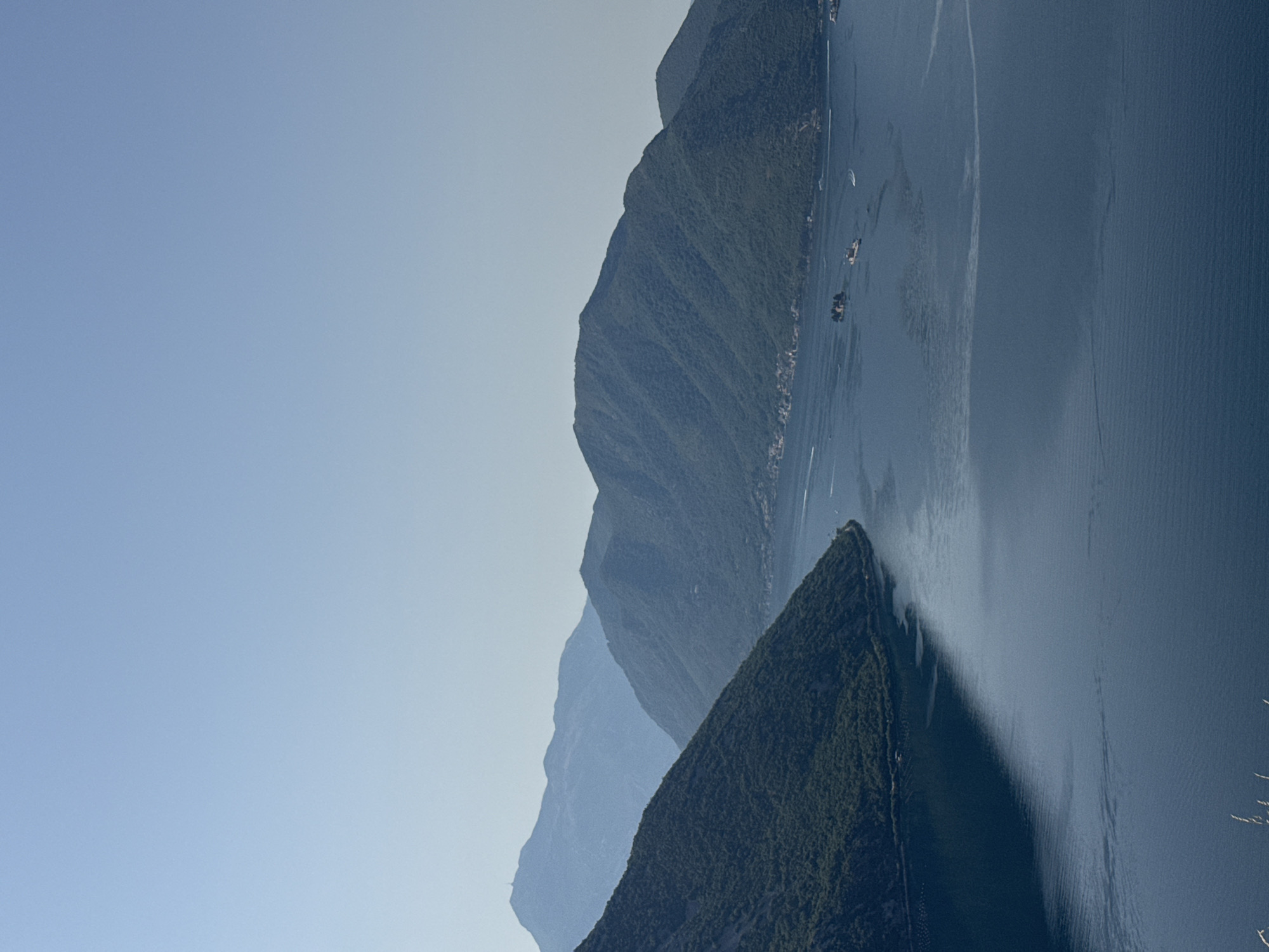 The image captures a panoramic view of a bay or fjord, likely the Bay of Kotor in Montenegro, with steep, verdant mountains rising from the water. In the foreground, a dark body of water reflects the blue sky and the silhouettes of the mountains. The water is rippled, suggesting a gentle breeze. Several boats, including what appears to be a larger vessel and smaller speedboats, are visible on the water, leaving faint wakes. Along the distant shoreline, nestled at the base of the mountains, small settlements with red-tiled roofs can be discerned. The lush green slopes of the mountains are dotted with trees, and in the far distance, several layers of mountains fade into a hazy blue. The sky is clear and a pale blue, transitioning to a lighter shade near the horizon. The overall scene evokes a sense of natural beauty and tranquility, characteristic of coastal mountain landscapes. The description is a general observation of the image and does not specifically place it in the town of Strp, Montenegro.