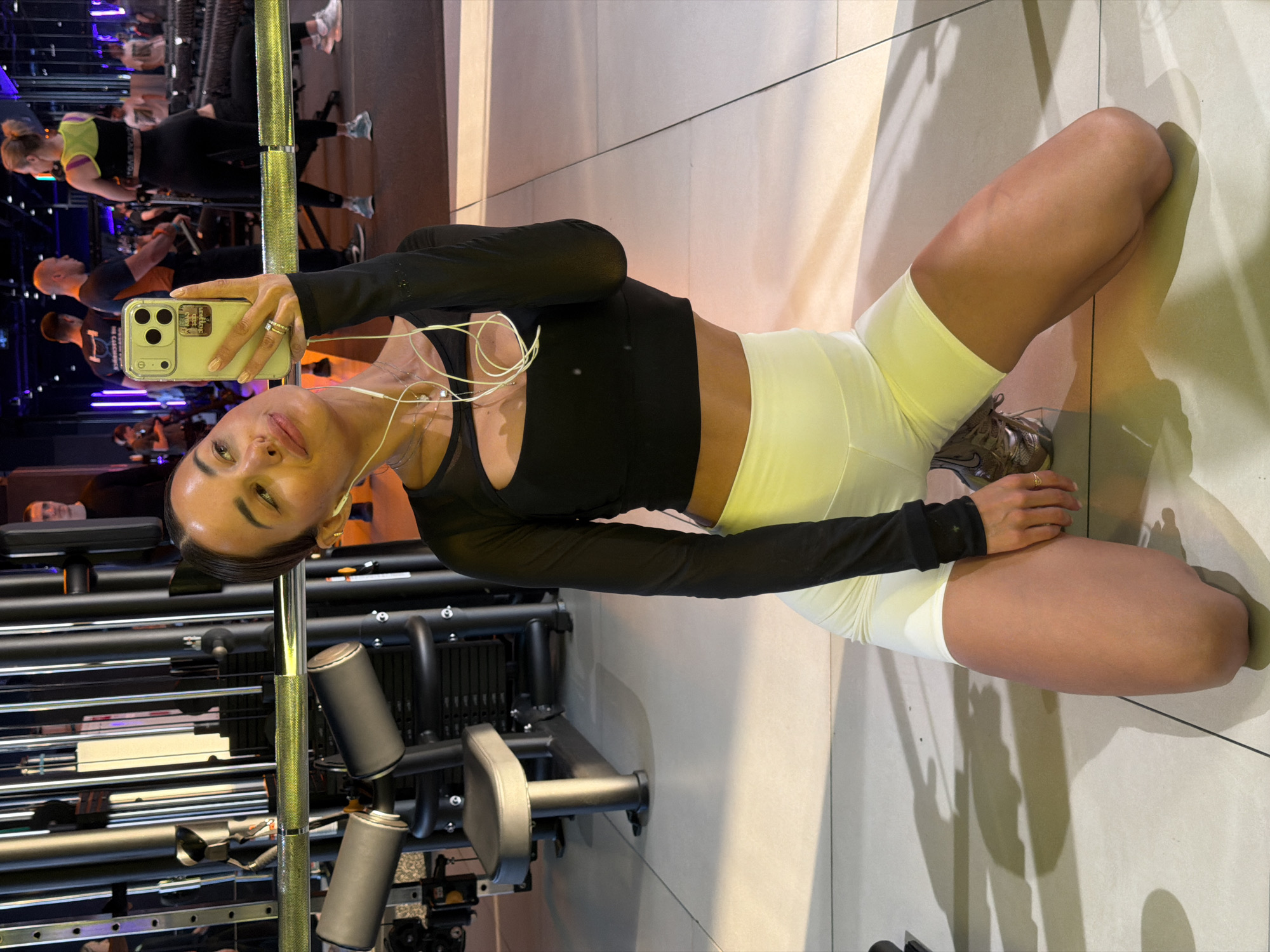 A woman is kneeling on a light-colored tile floor, taking a selfie with a smartphone. She is wearing a black long-sleeved athletic top and white shorts, with headphones around her neck. The background shows a gym environment with exercise equipment and other people working out. The exact location in Krasnoyarsk, Russia, cannot be determined from the image.