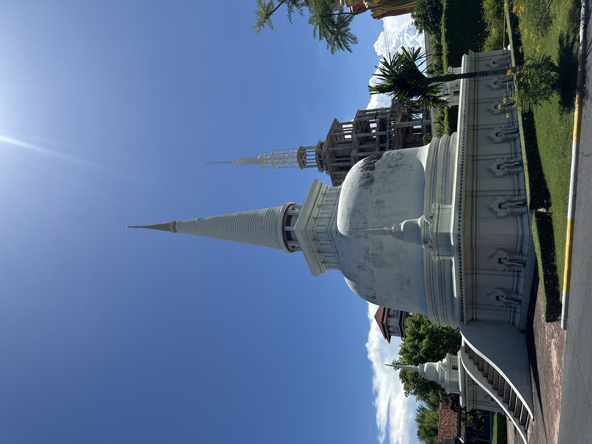 The image captures a white stupa, likely a Buddhist monument, under a bright blue sky in Bang Pu Subdistrict Municipality, Thailand. The stupa features a conical spire and a rounded base, with decorative elements along its lower sections. In the background, there is another structure appearing to be under construction, possibly another stupa or a building with a similar architectural style. The environment includes manicured grass and greenery, indicating a well-maintained area. The presence of sunlight and shadows suggests a daytime setting.