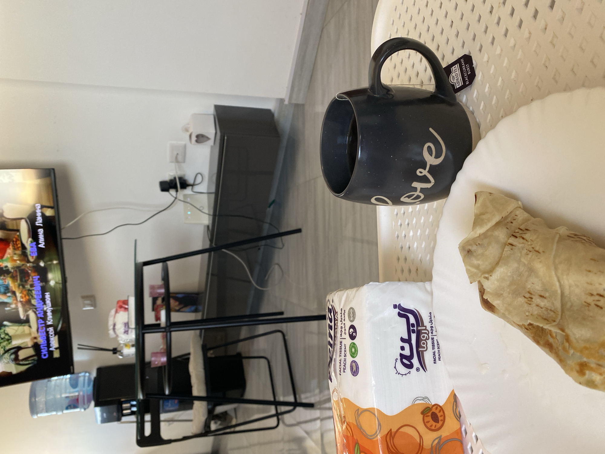 This media file captures a casual indoor scene, likely in Sharm El Sheikh, Egypt, given the Arabic text visible on the tissue box. The primary subjects are objects arranged on a white plastic chair. A dark mug with the word "love" inscribed in white cursive script sits to the left, containing a dark liquid, possibly coffee or tea. A tea bag tag from "AHMAD TEA" with "BLACKCURRANT BURST" is attached to the mug. To the right of the mug, on a white plate, is a wrapped food item, appearing to be a wrap or tortilla, partially unwrapped to reveal its filling. In front of the mug and plate is a package of "REINA" brand facial tissues, scented with peach, as indicated by the Arabic and English text "برائحة الخوخ" and "PEACH SCENT" respectively. The background is blurred but suggests a home environment with a television screen displaying text in Cyrillic script, possibly indicating a show or movie. Electrical outlets and wires are visible on the wall behind the television. A water dispenser and a black metal shelf unit with some items on it are also present in the background, adding to the domestic setting. The lighting suggests daytime. The overall impression is that of a relaxed moment, perhaps breakfast or a snack break.
