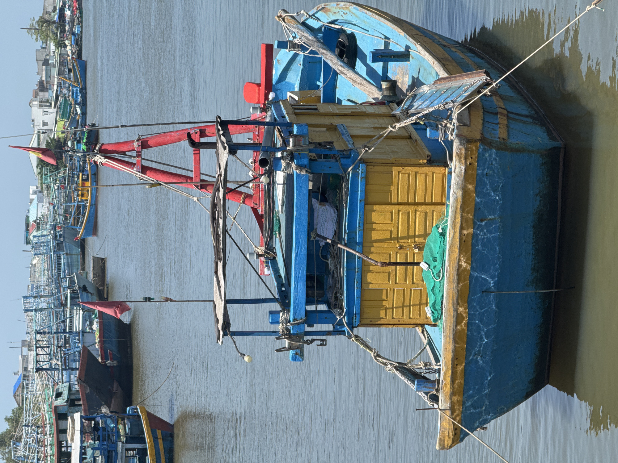 The image depicts a fleet of colorful fishing boats docked in a harbor. The foreground is dominated by a prominent blue and yellow fishing boat with a wooden superstructure. The boat features a red A-frame structure with a small Vietnamese flag attached. Ropes and fishing nets are visible on the deck. In the background, numerous other fishing vessels of varying sizes and colors are moored together. The water is calm and reflects the sky. The scene appears to be located in a coastal urban area, likely Phu Thuy, Vietnam, with buildings and trees visible on the shore in the distance. The sky is clear and the lighting suggests it is daytime. There are no people visible in the image.
