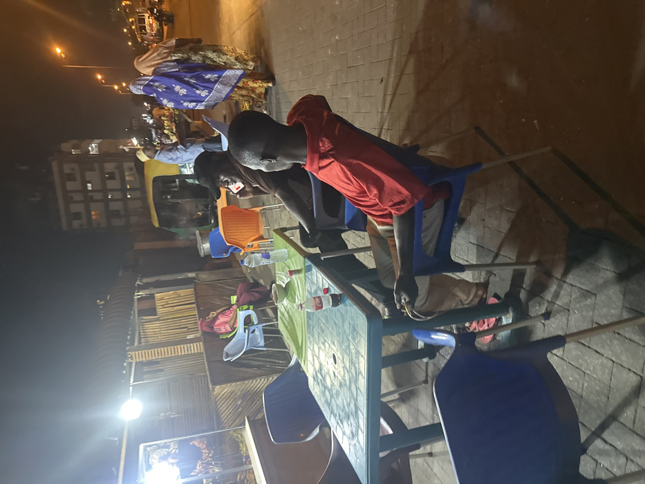 At night, a person in a red shirt sits at a green plastic table, holding a yellow object. Another person is seated next to them, looking at a phone. Blue plastic chairs surround the table. A small building constructed from corrugated metal is visible in the background, along with street lights, pedestrians, and parked vehicles. The ground is paved, casting long shadows from the table and chairs. Bottles of liquid are on the table.