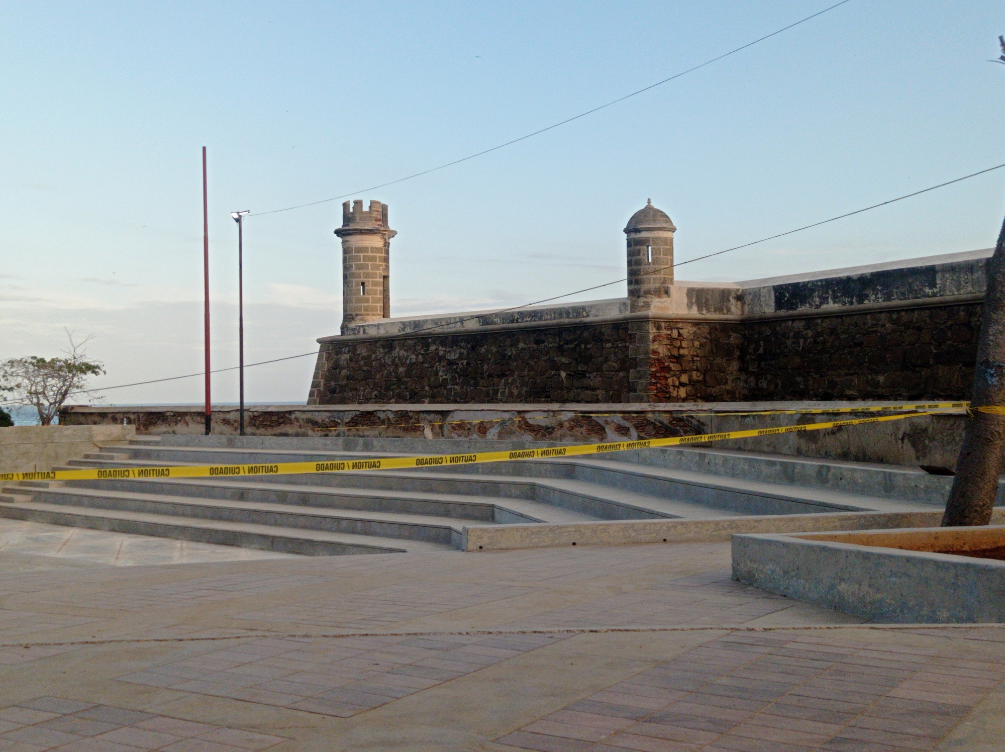 The image captures a view of the Castillo San Carlos Borromeo in Pampatar, Venezuela, a historic fort. The foreground presents a paved square leading to a series of wide, shallow steps, partly blocked by yellow caution tape, bearing the text "CAUTION" in mirror image. The steps ascend to the base of the fort, which is constructed of dark stone and features two rounded turrets at the corners. Above the fort wall, a clear, light blue sky suggests either early morning or late afternoon light. Two tall, thin poles are visible on the left, adding vertical elements to the composition. A bare tree stands to the left of the steps, and a segment of a tree trunk is visible to the right.