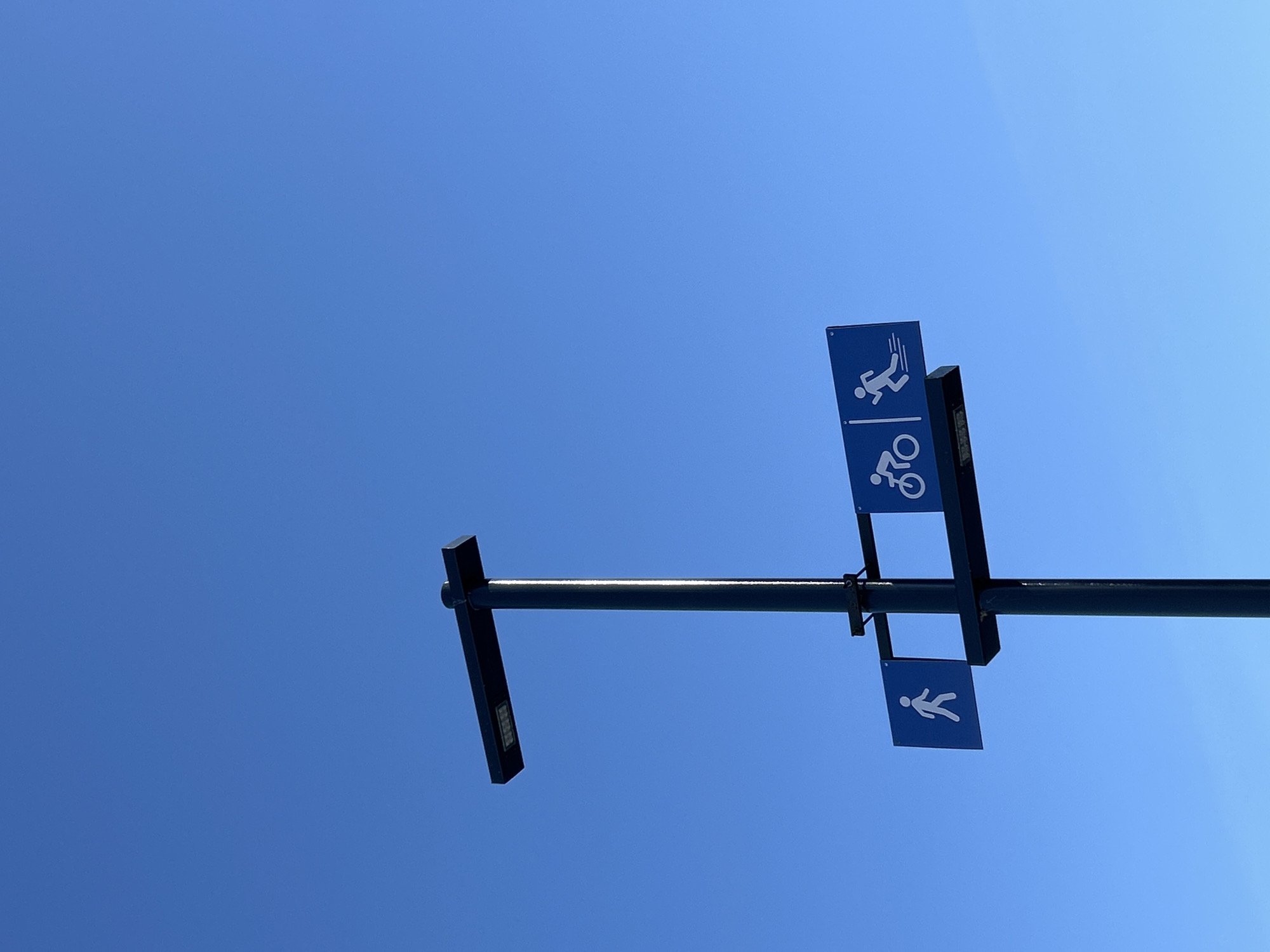 The image presents a minimalist view of a clear, bright blue sky, indicating a sunny daytime setting. Dominating the upper right quadrant is a dark, horizontal pole or crossbar, likely metal, extending across the frame with a slight upward tilt. Attached to this pole are two distinct blue rectangular signs featuring white pictograms.
The upper sign, positioned more centrally, displays two symbols separated by a horizontal line: a person running or jogging at the top, and a bicycle below. This arrangement typically denotes a shared route or distinct lanes for both runners and cyclists. Further down and to the right, a second, smaller blue sign is visible, depicting a single white pictogram of a person walking. This suggests a designated path for pedestrians.
The horizontal pole has darker, rectangular extensions at its ends; the left extension shows a textured surface that might be a luminaire or a decorative element. The sharp focus on the signs against the expanse of the unblemished sky highlights their informational purpose. There is no visible text on the signs, only the universally recognized pictograms. The scene is devoid of people or other objects, emphasizing the signs and the vast, clear sky. This type of signage is common in outdoor recreational areas or urban infrastructure, such as those found in Chaikovsky, Russia, the specified location.
