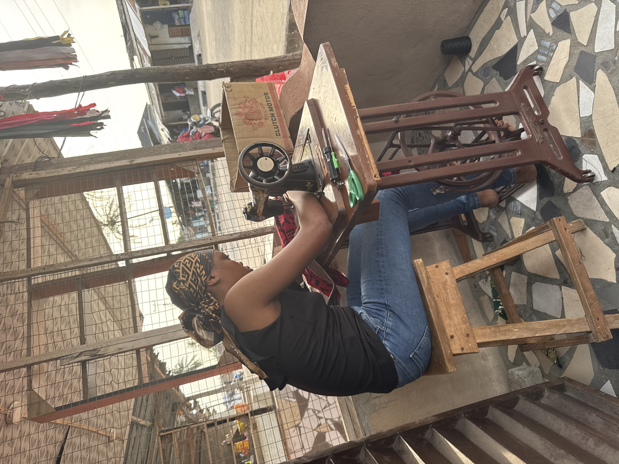 An outdoor scene in Badagry, Nigeria, shows a woman seated at a vintage sewing machine. She wears a black tank top, blue jeans, and a headscarf. The sewing machine, with a “Clutch Motor” box behind it, is set up on a wooden table with a metal frame. The environment is a mix of metal fencing, concrete structures, and patterned tile flooring. There are tools hanging on the sewing machine table. A roll of thread is visible on the floor. Clothes hang on a line in the background. The scene is open-air, with various objects and structures suggesting an urban setting.