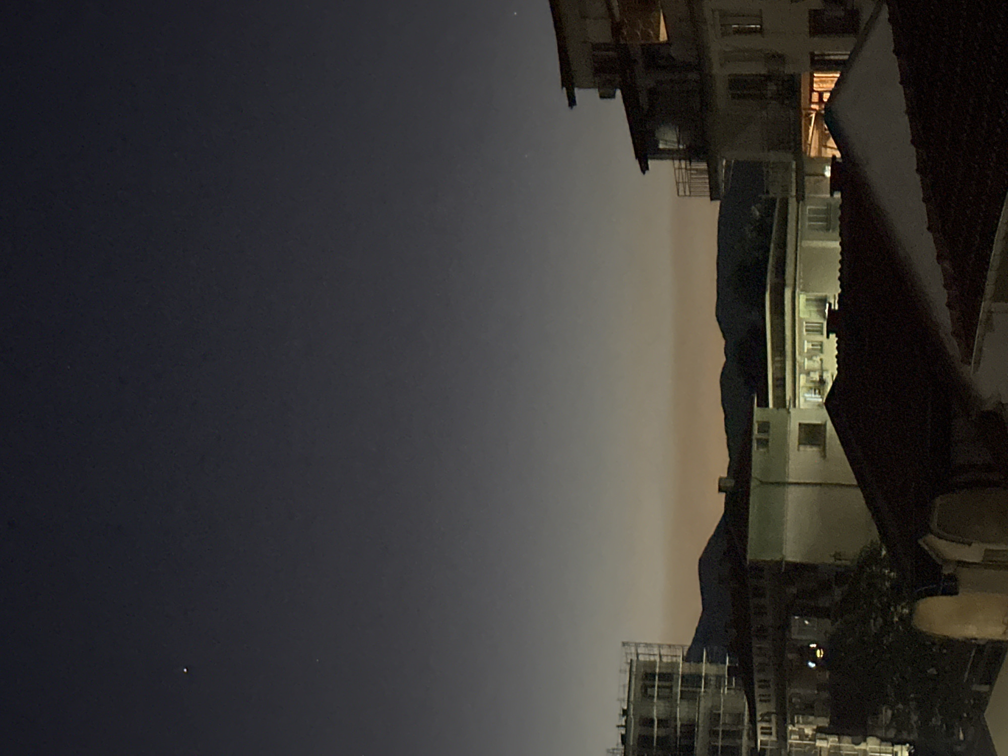 The image depicts a nighttime cityscape. The sky transitions from dark to light, suggesting either twilight or light pollution. Buildings of varying heights are visible, some appearing old and some modern. The presence of mountains can be discerned in the distance. The location is unspecified and cannot be determined from the image alone. The scene appears still and devoid of visible human activity.