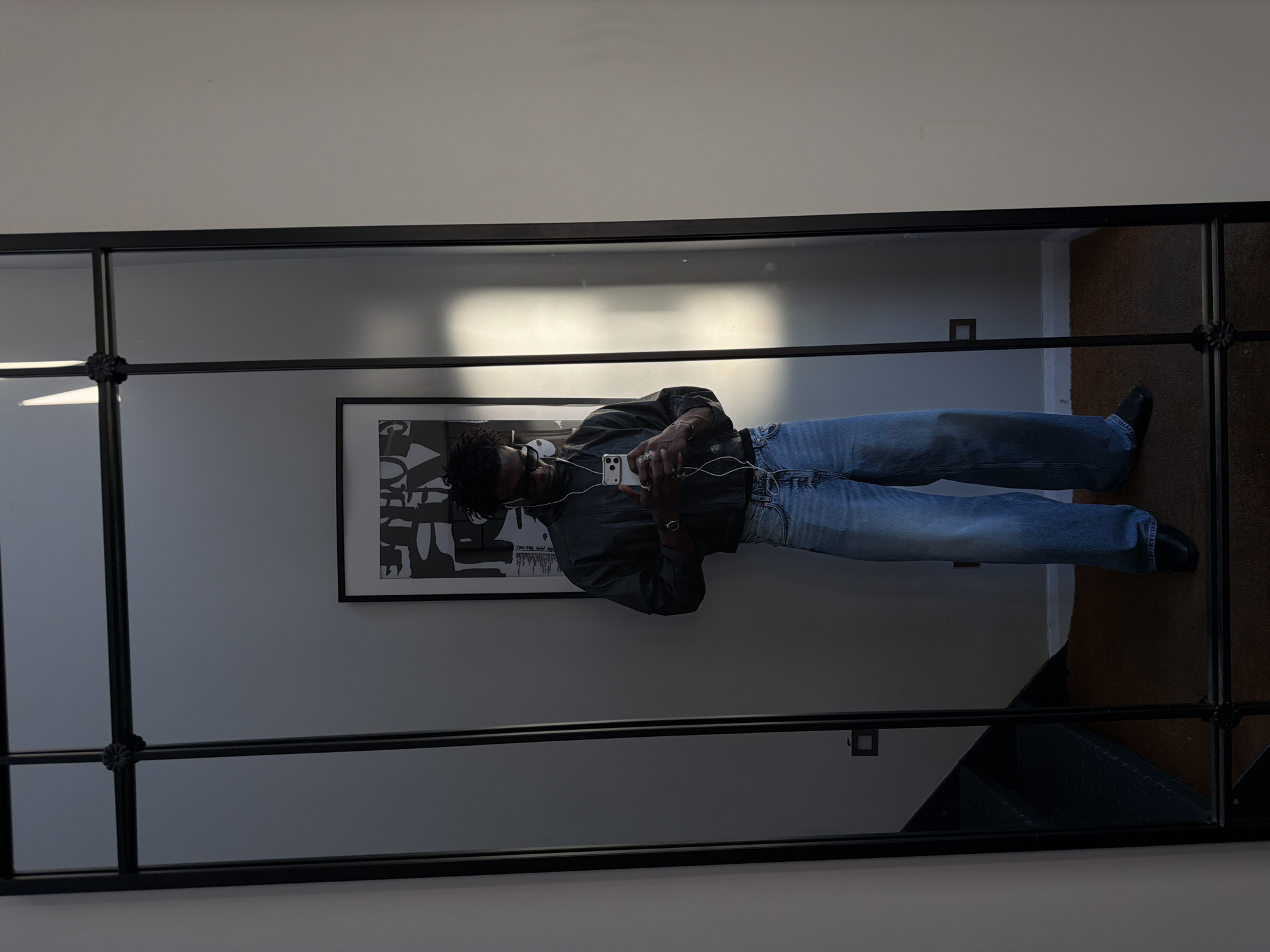 A full-length mirror reflects a man standing in a room, taking a selfie with his phone. He is wearing a dark, high-necked shirt and light-wash, wide-leg jeans. White earbuds are visible, connected to his phone. He has dark hair and is wearing sunglasses. In the background of the reflection, a piece of abstract art hangs on the wall, and a staircase is visible to the left. The room appears to be an apartment or house, with neutral-colored walls and a rug on the floor. The lighting suggests it is daytime, with sunlight casting a shadow on the wall. The man's pose and the presence of a smartphone indicate he is engaged in taking a photograph. The style of the mirror and artwork suggests a modern, urban interior design, possibly in London, United Kingdom, though no specific landmarks or indicators confirm this location.