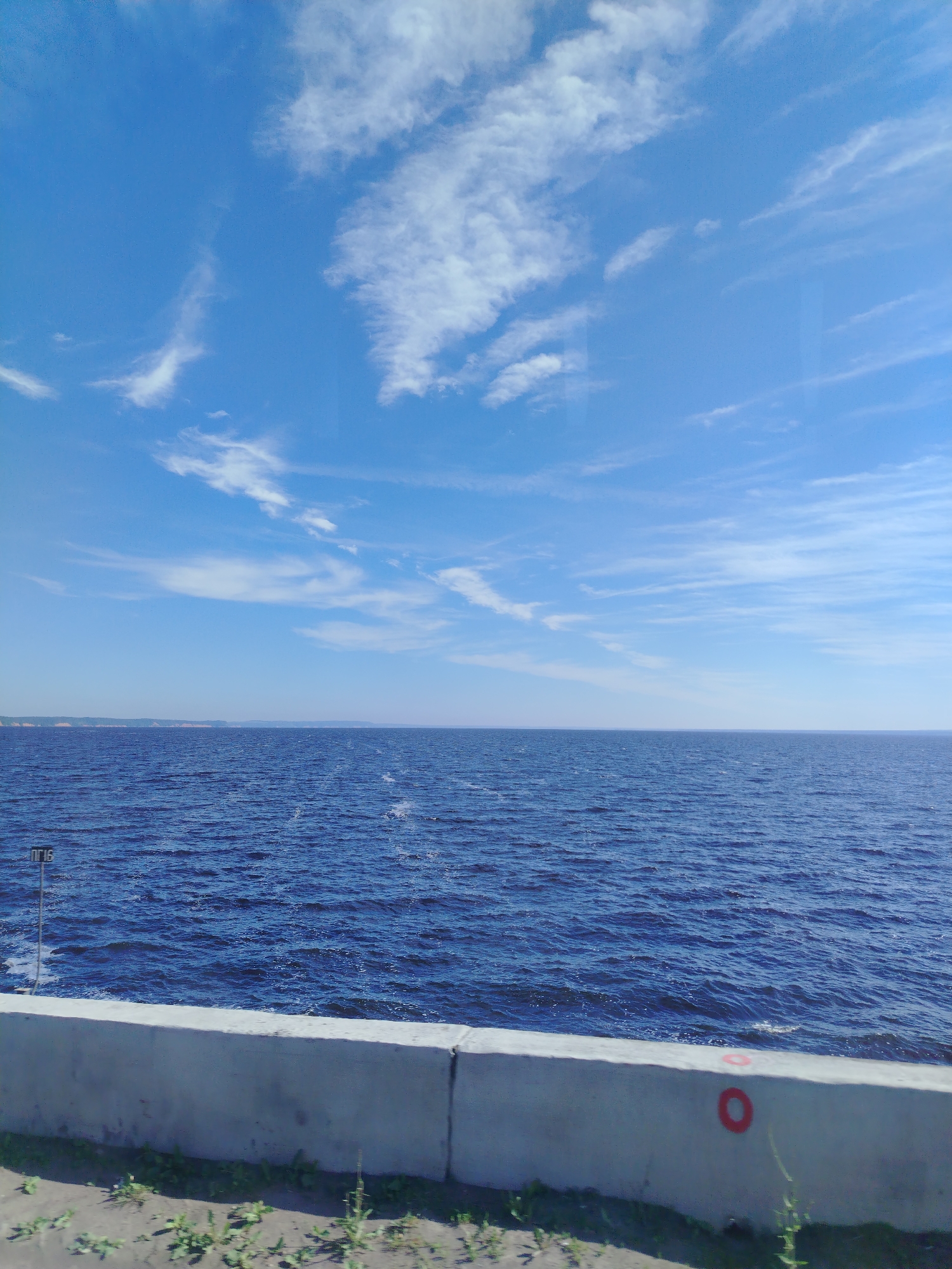 The media file presents a scenic landscape dominated by a vast body of deep blue water under a bright blue sky. The image appears to be taken from an elevated position alongside the water, possibly a road or embankment.
In the foreground, a light gray concrete barrier or railing runs horizontally across the bottom, separating a strip of dry, sparse green grass and dirt from the water. On the left side of this barrier, a thin metal pole rises from the water or bank, topped with a white sign displaying the Cyrillic text "ПГ16". Towards the right, a distinctive red circular marking is painted on the concrete barrier.
The mid-ground is filled with the expansive, deep blue water, showing gentle ripples on its surface. In the far distance, a faint, low-lying landmass or coastline with subtle hills or cliffs is visible, stretching across the horizon.
Above the water and land, the sky is a vibrant, clear blue, adorned with scattered, wispy white clouds that extend across the entire upper portion of the frame. These clouds appear to be cirrus or cirrocumulus, indicating fair weather.
There are no people or specific activities taking place, creating a sense of tranquility and openness. The bright lighting suggests it is daytime, likely late morning or early afternoon. Given the location context of Chaikovsky, Russia, and the Cyrillic text, this vast body of water is likely the Kama Reservoir.