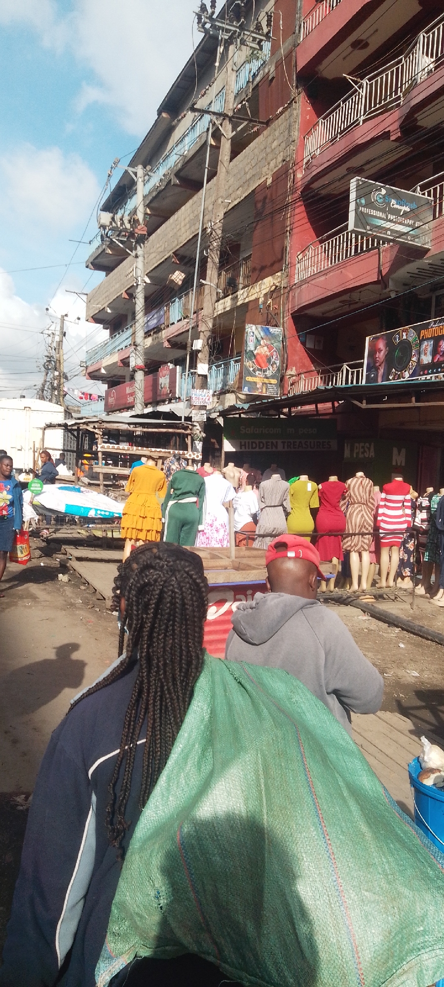 The image shows a bustling street scene in Nairobi, Kenya. In the foreground, two individuals stand with their backs to the camera: a person with braided hair wearing a blue jacket and a man in a grey hoodie and a red cap. A large green bag rests on their shoulders.
Behind them, a row of mannequins displays various articles of clothing including dresses and sportswear. The mannequins are positioned in front of a series of storefronts, under signs. One sign reads "HIDDEN TREASURES", with the text "Safaricom m pesa" appearing above it. Other signs are visible, including one indicating a photography studio.
In the background, a multi-story building with several balconies looms over the street. Electrical wires crisscross above the street. People are moving about in the street. The overall impression is of a crowded, active commercial area.