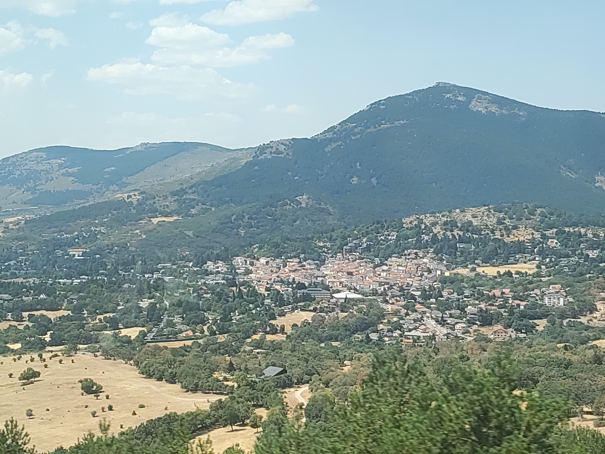 This is an aerial, eye-level, wide shot of Navacerrada, Spain. The scene features a village nestled in a valley with lush green trees and brown fields. A large mountain range dominates the background, with a clear blue sky and scattered clouds above. The village has a dense cluster of buildings with red-tiled roofs, suggesting a traditional Spanish architectural style. Some larger, modern buildings are also visible. The foreground is filled with dense trees, partially obscuring the view. The image appears to be taken on a sunny day, with the sun casting shadows on the landscape. There are no people or animals visible in the image. The overall impression is a serene, rural landscape with a well-established village.