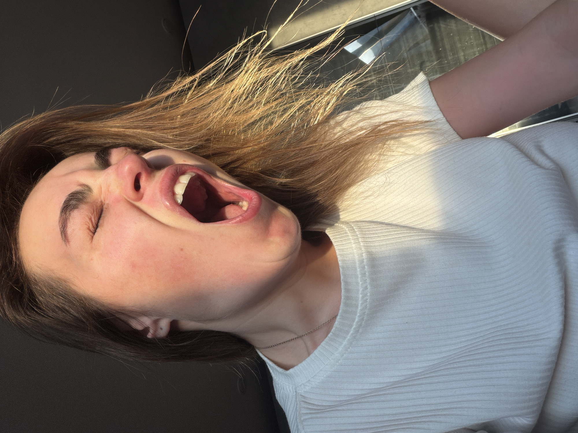 A woman with light brown hair, closed eyes, and an open mouth, possibly yawning or shouting, is the primary subject. She is wearing a white, ribbed shirt and a delicate silver necklace. The scene is illuminated by bright sunlight, which highlights the hair and casts shadows on the face. The background is blurred and includes parts of a dark grey interior, possibly a car, and a window showing a blurry exterior. No specific details connect the image to the city of Izhevsk, Russia.