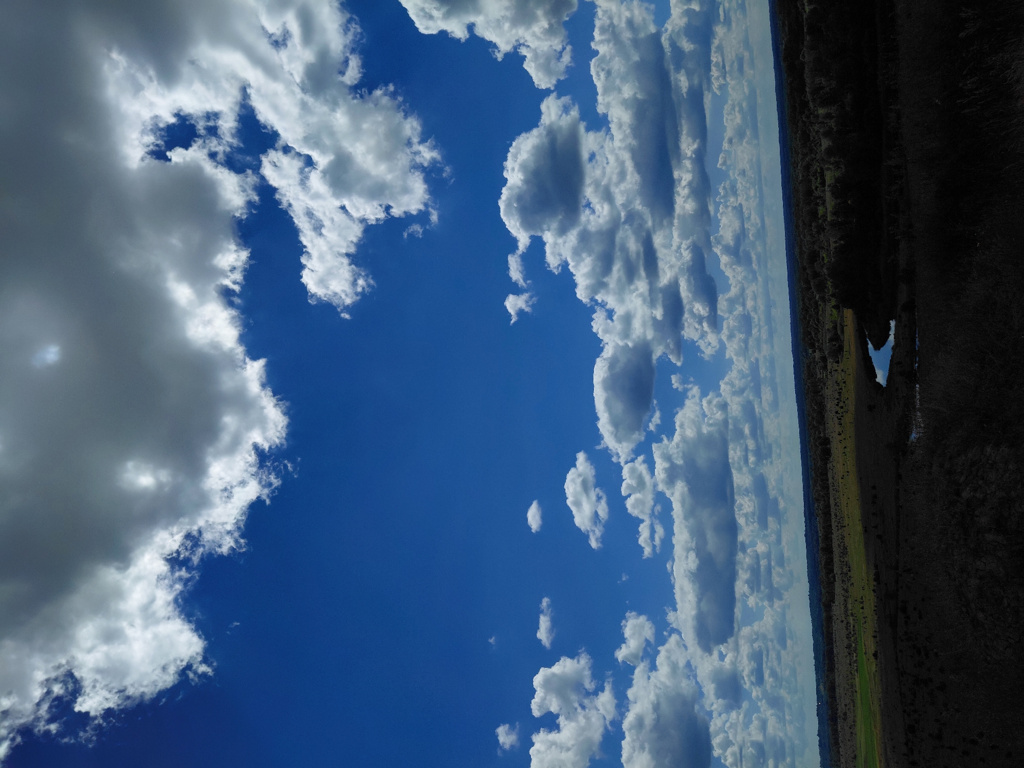 This is a landscape photograph taken in Ustyvytsia, Ukraine, under a bright blue sky filled with scattered white and gray clouds. The foreground is a dark, shadowed area with indistinct vegetation. A winding, reflective body of water, likely a river or stream, is visible in the mid-ground, curving through a landscape of fields and scattered trees. The horizon is marked by a line of trees and undulating terrain, extending into the distance under the expansive sky.