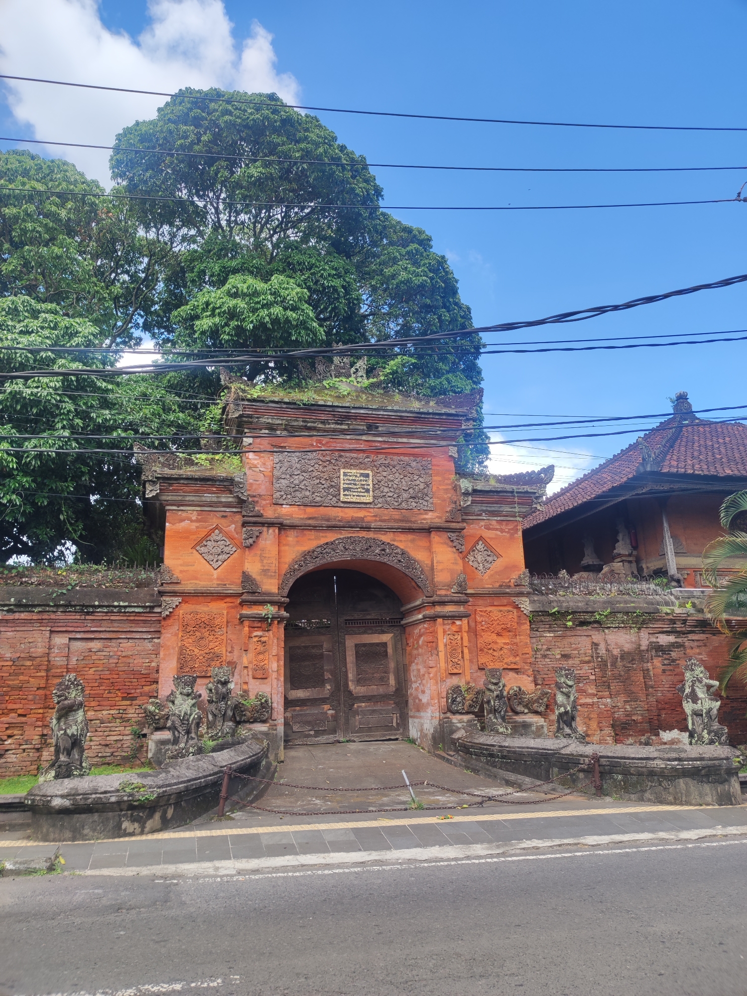 The image shows a traditional Balinese gate in Gianyar, Indonesia. The gate is made of red brick and stone, with intricate carvings and sculptures. There are several statues of mythical creatures flanking the gate's entrance. The gate is closed with large wooden doors and a chain. Vegetation covers the gate and surrounding structure, indicating its age. A building with a traditional Balinese roof is visible to the right of the gate. Power lines run across the sky above the gate and trees. The street is visible in the foreground. The sky is blue with scattered clouds.