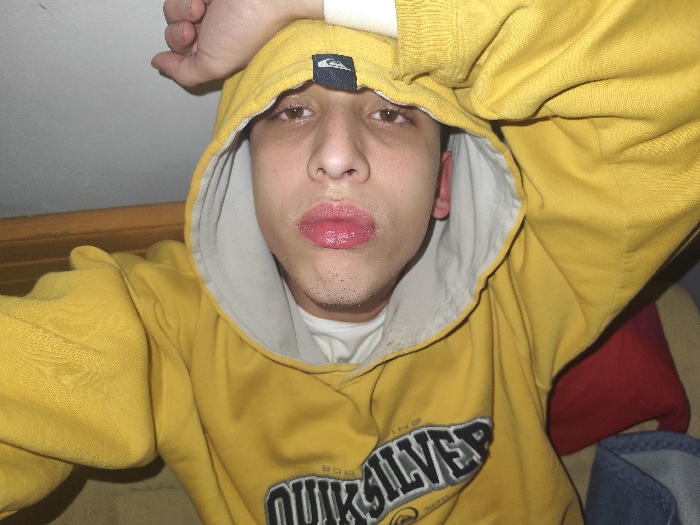 A person with light skin is centered, wearing a yellow Quiksilver hoodie. The hood is up, framing their face. Their lips are noticeably glossy. One hand rests on top of the hood. A partial view of a wooden headboard is in the background to the left, while a red pillow and blue denim appear on the right. The person is looking directly at the viewer with a neutral expression.