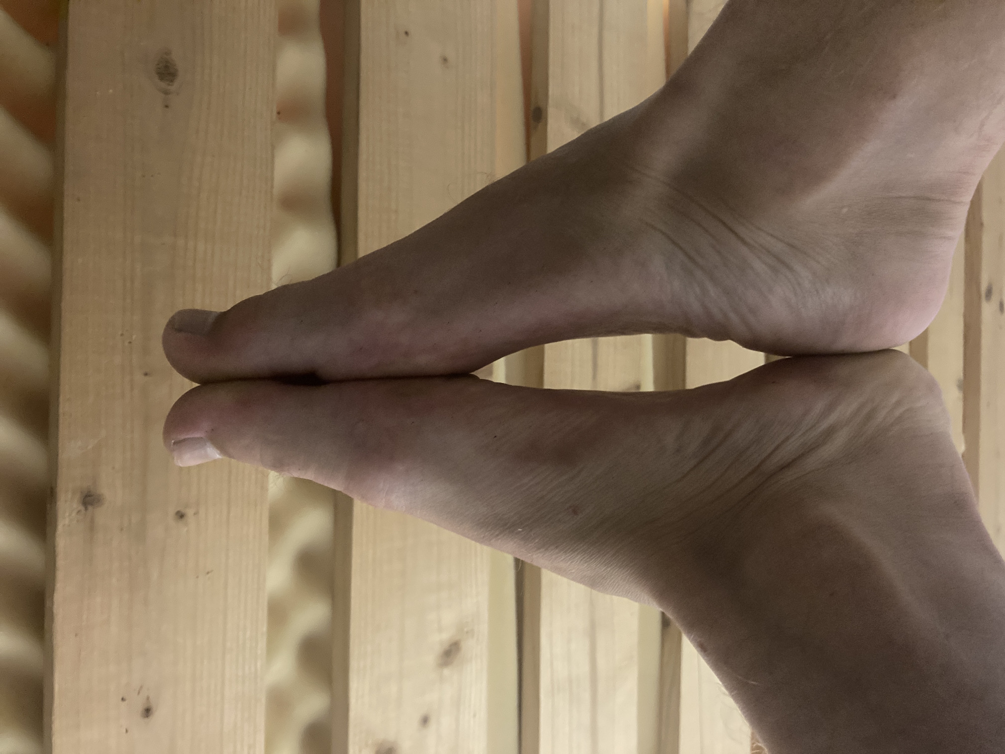 The image displays a close-up of a bare human foot and its reflection, creating the appearance of two feet. The upper foot rests with its toes on the sole of the lower, mirrored foot, forming a symmetrical, almost triangular shape. Both feet have a light skin tone, and details such as toenails, skin texture, and fine hairs are visible.
The feet are positioned against a background of vertical wooden planks, which are light brown or beige with visible grain and knots. The surface on which the feet are resting is highly reflective, creating a clear mirror image of the lower foot. The lighting in the scene appears warm, likely from an indoor artificial source, contributing to the slightly yellowish hue of the wood and skin. There are no discernible shadows that would indicate strong directional natural light. The indoor setting, with its wooden paneling, in Vessigebro, Sweden, could suggest an environment such as a sauna or a room with wooden interior finishes. No text is visible in the image.