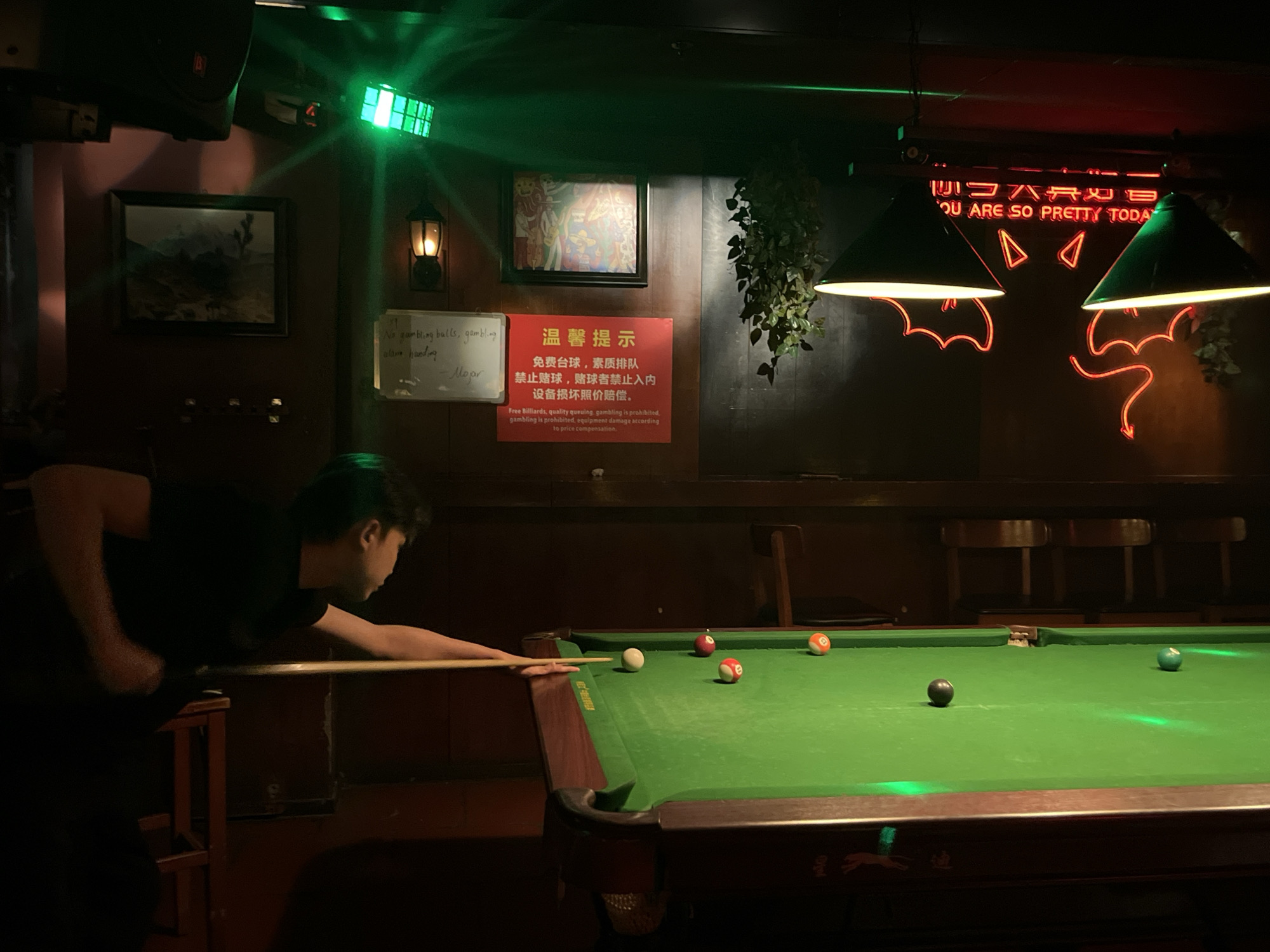 A dimly lit interior scene shows a person, likely in their late teens or early twenties, playing pool on a green-felted pool table. The person is leaning over the table, cue in hand, preparing to strike the cue ball. Several colored pool balls are scattered on the table. Above the table, two lamps with cone-shaped shades hang from the ceiling. Behind the table is a dark wall decorated with various items, including framed artwork, a cautionary sign in Chinese and English, and a neon sign in Hebrew, which translates to "You are so pretty today." A small white board with a handwritten note is also visible on the wall. Based on the text on the warning sign, this image was likely taken in Liangjiang, China.