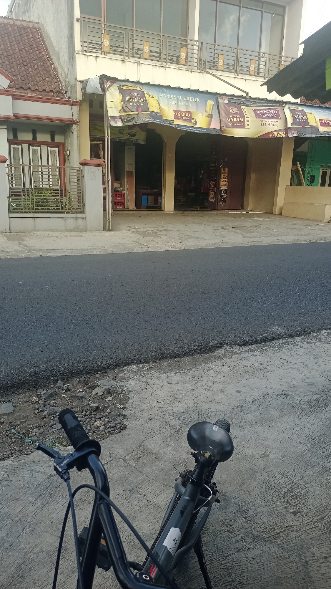 The image shows a street view in Pakemitan, Indonesia, featuring a commercial building with a banner displaying advertisements for kretek cigarettes. In the foreground, a black bicycle is parked on a concrete sidewalk. The building has a tiled roof and multiple windows. The banner on the building is yellow and features text and images related to cigarette brands like "Surya Nusantara Rava" and "Gudang Garam". The street in front of the building appears to be paved. The scene is likely captured during daylight hours.