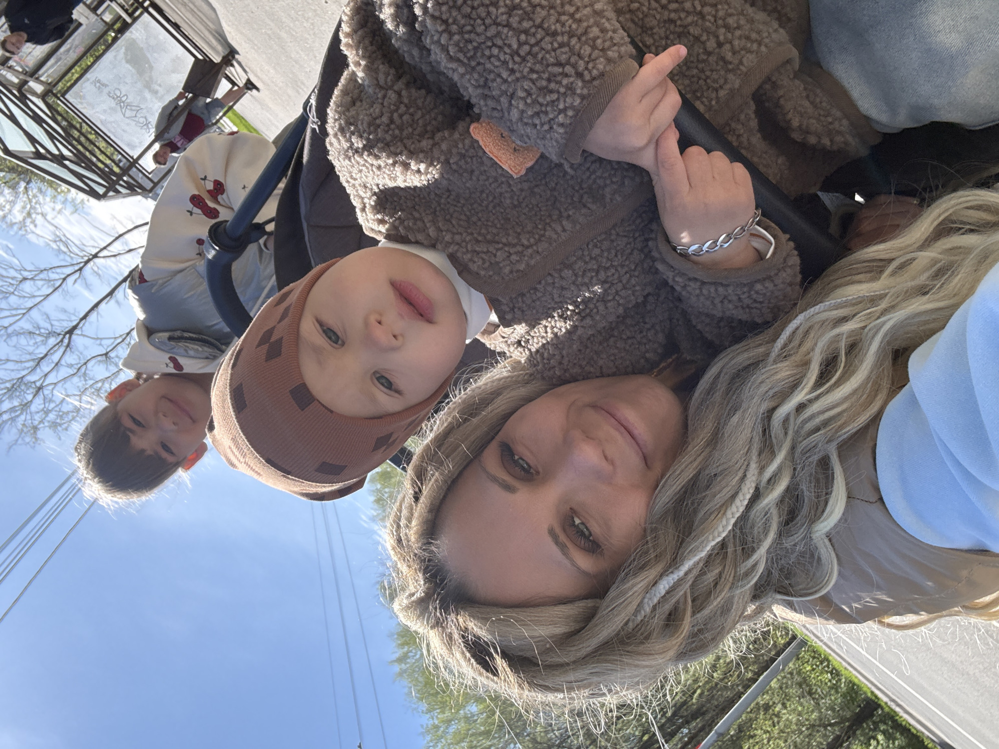 A woman is taking a selfie with two children. The younger child is sitting in a stroller. The older child is standing next to the stroller. The background shows a blue sky, trees, and utility poles. The location is presumably somewhere in Rusanovo, Russia.