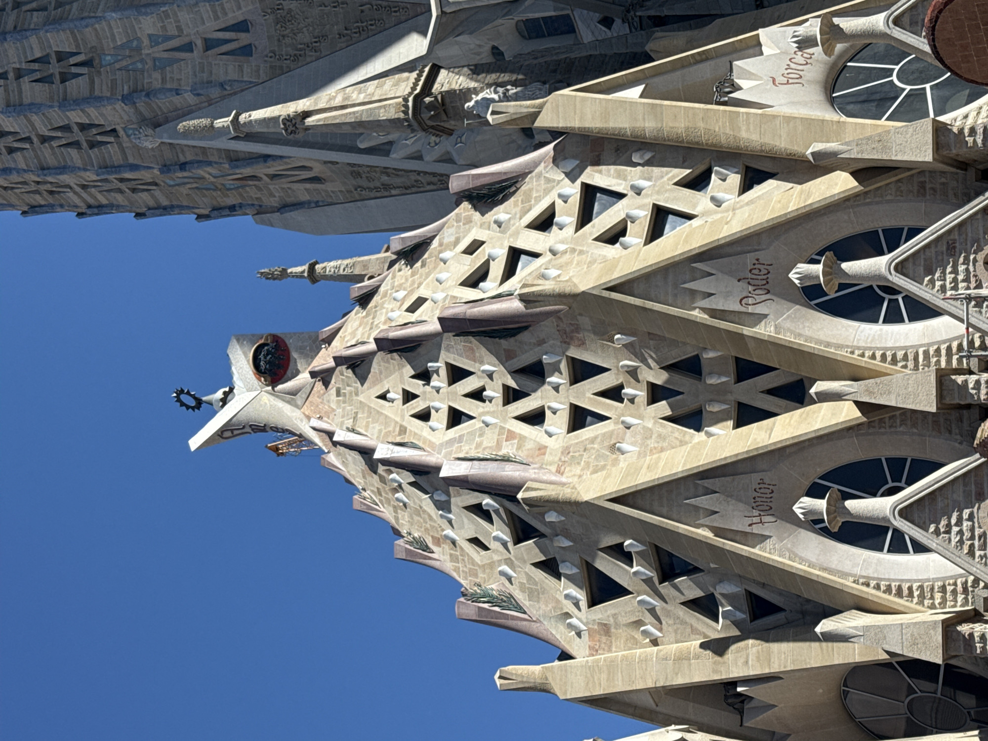 The image depicts the upper portion of the Sagrada Familia basilica in Barcelona, Spain, angled against a clear blue sky. The architectural details are prominent, showing the intricate stonework and design elements, including triangular windows and decorative inscriptions like "Honor", "Poder" and "Força." A decorative star or pinnacle is visible against the sky near the top left. The stone appears light in color.