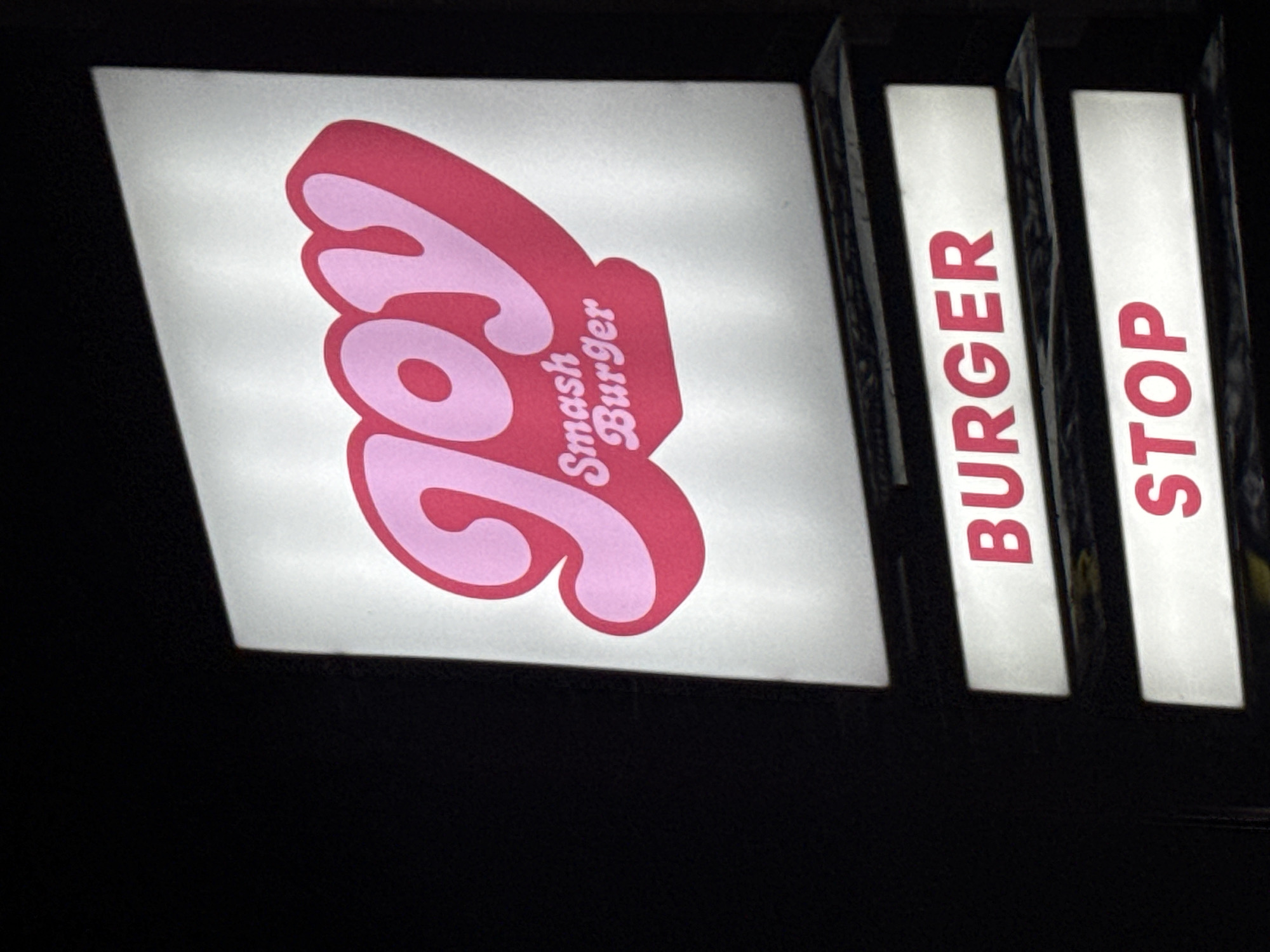 The image shows a sign for "Joy Smash Burger Stop" in North Kuta, Indonesia. The sign is illuminated with a pink retro font logo and a white background. To the right of the logo are two vertical rectangular signs, one displaying "BURGER" and the other "STOP" in red lettering. The background is dark and the signs are brightly lit.