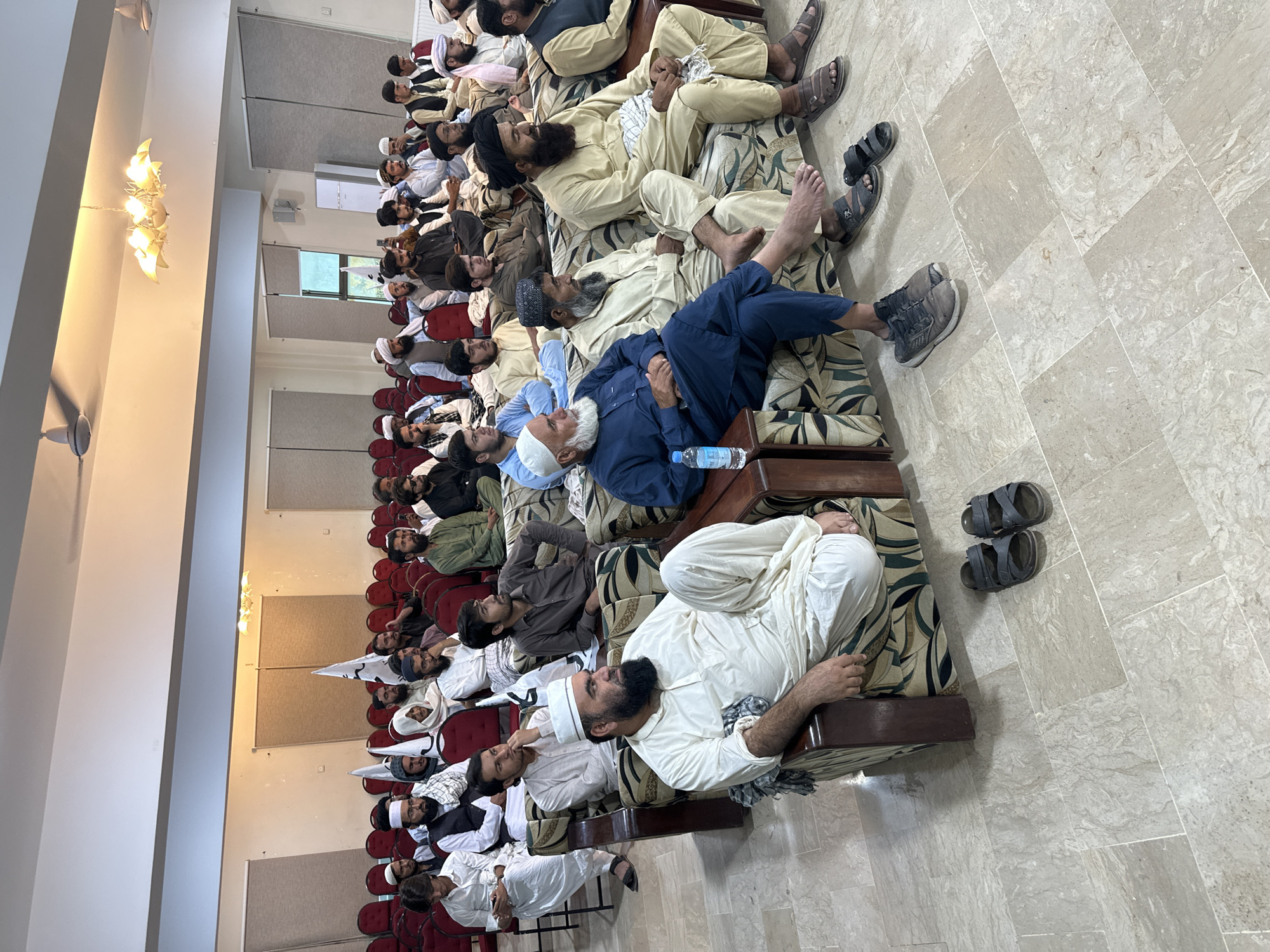 The image depicts a gathering of men lying on cushioned benches in what appears to be an indoor meeting hall. The location is Quetta City Tehsil, Pakistan. The men are predominantly dressed in traditional Pakistani clothing, including shalwar kameez and various head coverings. Some of the men are barefoot or wearing sandals, while at least one wears sneakers. Many are lying down, seemingly resting or relaxing. A water bottle is visible next to one individual. In the background, rows of chairs line the wall. The floor is tiled. The environment is well-lit with a large chandelier visible in the upper left of the frame.