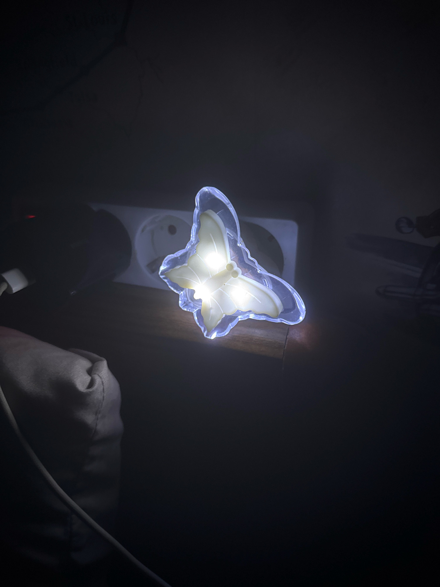 A glowing butterfly-shaped night light is plugged into a wall socket. The night light is translucent with white and yellow coloring. The socket is recessed into the wall. To the left of the socket, a white electrical cord is visible, connected to an unseen device. In the foreground, a beige fabric object, possibly a bag, is partially visible, with a curved wire resting on it. The background is dark and appears to be an interior setting. The image was taken in a location in the city of Hrodna, Belarus.