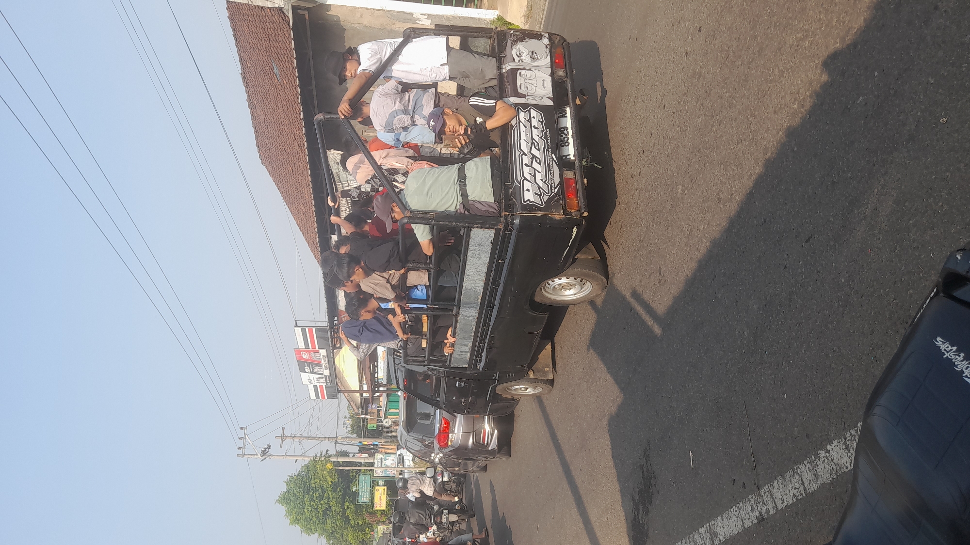 A black pick-up truck, its rear bed filled with passengers, drives along a paved road in Lemahabang, Indonesia. The truck is adorned with stickers on its rear, one featuring cartoonish drawings of faces and text. A license plate on the truck reads "R 8529 AM". Multiple people, including men, women, and children, are seated and standing in the open bed of the truck, some holding onto the side rails for stability. The truck is in motion, with other vehicles, including cars and motorcycles, visible in the background and on the side of the road. The environment appears to be a suburban or semi-urban area, with buildings, power lines, and trees lining the street. The sky is clear and a bright blue, indicating a sunny day. The overall scene depicts a common mode of transportation in the region, with a significant number of people being transported in the pick-up truck.