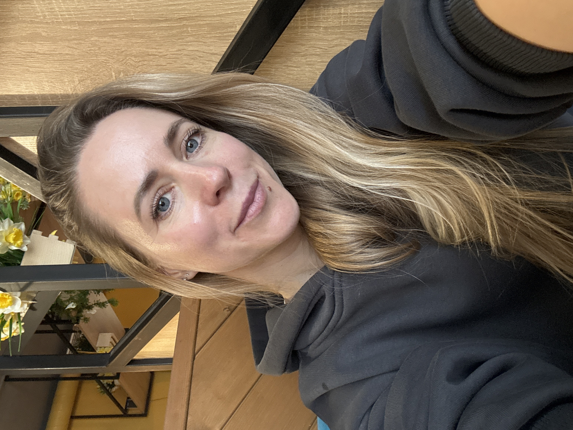 A woman with blonde hair and blue eyes wearing a gray hoodie is in an interior setting. A table is partially visible in the background. In the left background is a black metallic structure and some flowers. The location in the city of Chaikovsky, Russia, is not determinable from the visual information provided.