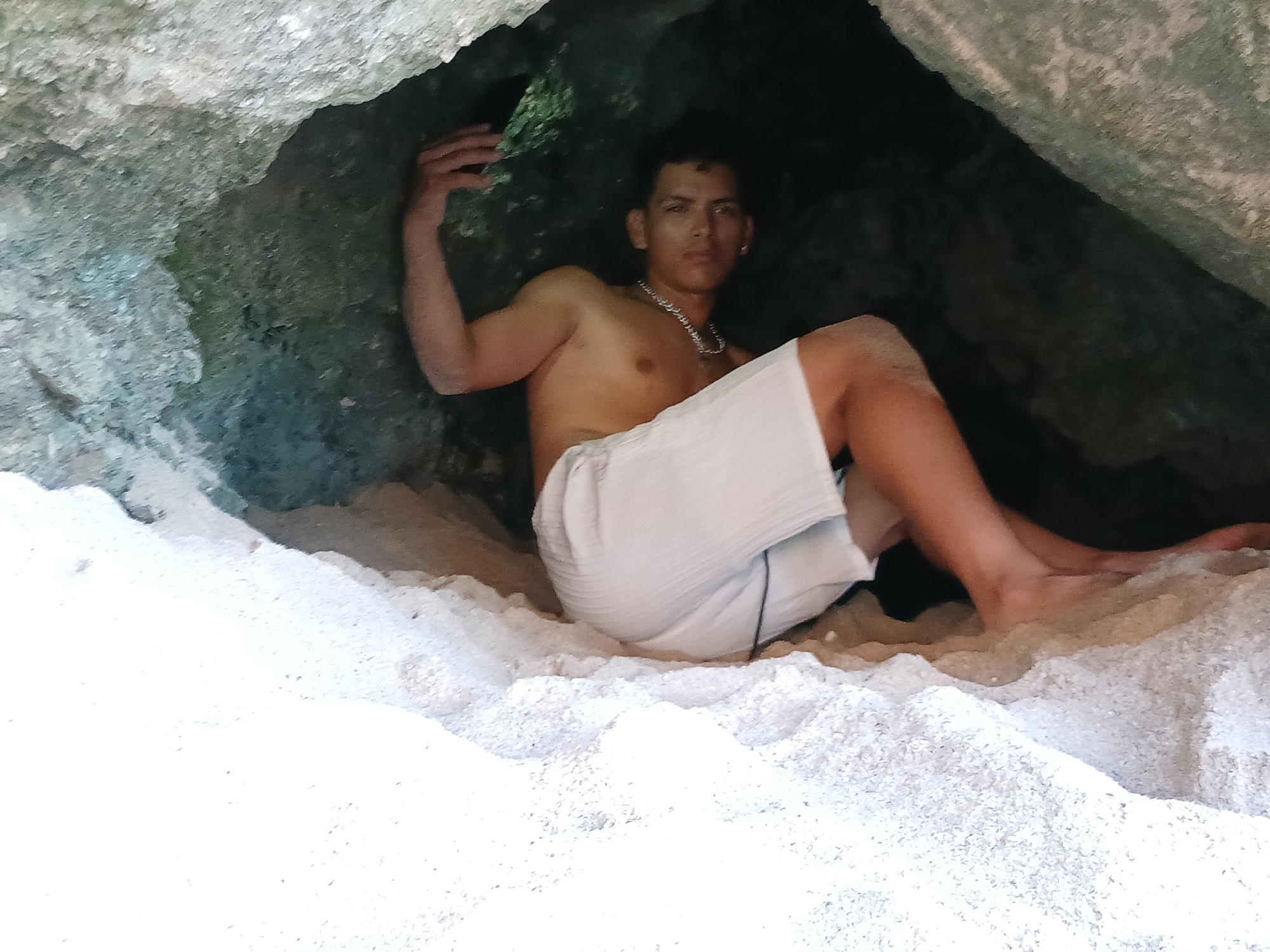 A young man is depicted sitting or reclining inside a natural cave or rock formation. He is shirtless, wearing white shorts, a silver chain necklace, and an earring in his left ear. His left arm is raised, with his hand resting on the cave wall, while he looks directly towards the viewer with a neutral expression.
The setting is characterized by very white, fine sand covering the ground, resembling soft beach sand. The cave walls are dark, rocky, and appear to have some natural texture, possibly moss or dampness in places, suggesting an organic formation. Natural light illuminates the sandy foreground, indicating it is daytime. The man's light attire suggests warm weather. There is no visible text in the image. Given the context, this scene is located in Juan Griego, Venezuela, likely a coastal cave or grotto.