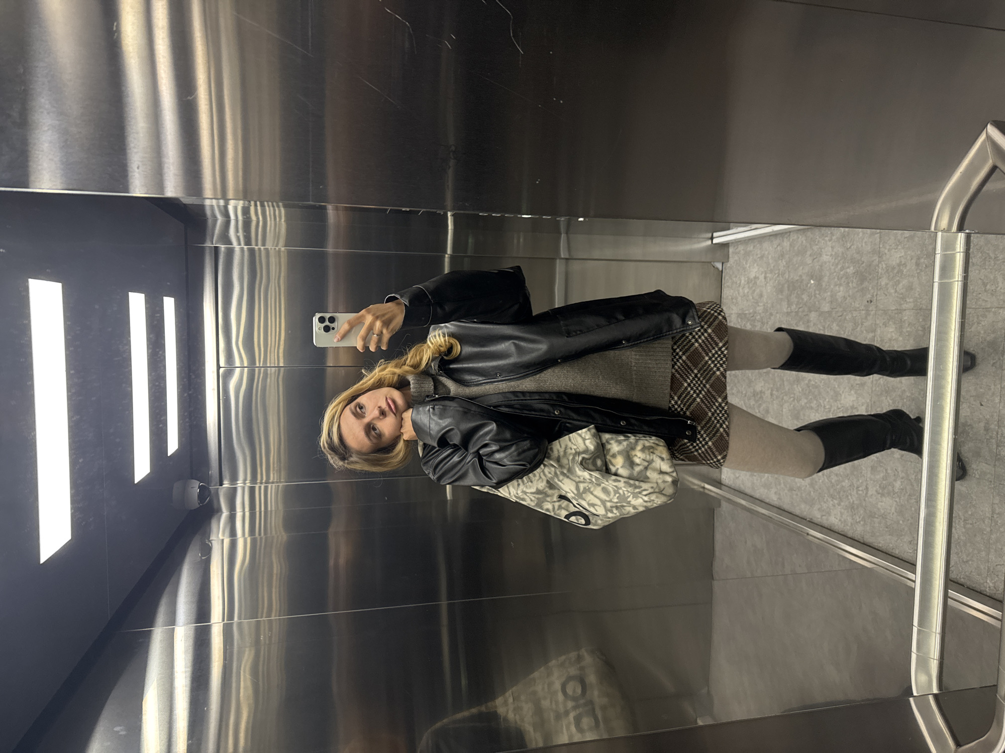 The image shows a person taking a mirror selfie inside an elevator. The subject, a woman with long blonde hair, is visible from the waist up, looking towards the camera while holding a light-colored smartphone in her right hand. She is wearing a black leather jacket over a grey top, a brown and white plaid skirt, light-colored tights, and black knee-high boots. A patterned, light-toned bag, whose design is not clearly legible, is carried on her left arm. A ring is visible on her right hand.
The setting is an elevator with highly reflective, metallic walls, displaying vertical streaking and reflections of the internal lighting. The floor is a light grey tiled pattern. On the left side of the elevator, multiple vertical strip lights illuminate the space, and their reflections are visible on the opposite wall. A small, white security camera is mounted in the upper left corner of the elevator's interior. A metallic handrail runs along the right wall. The lighting is artificial, creating bright highlights on the polished surfaces. The woman's expression is neutral, indicative of posing for a photograph. No discernible text is visible on any objects or surfaces within the image.