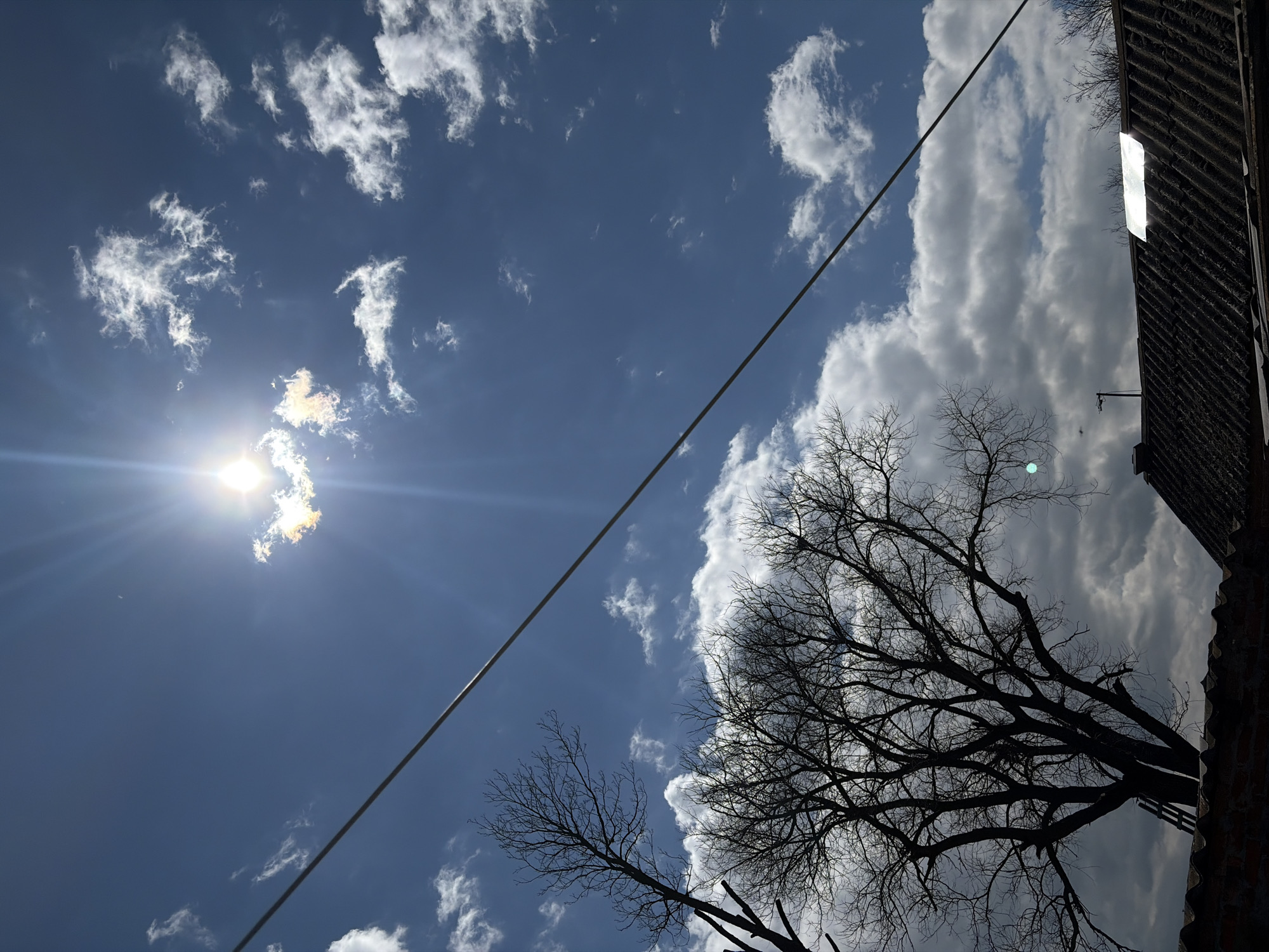 The image is a low-angle shot of a sky with scattered clouds, a bright sun, and a few bare trees silhouetted against the sky. A power line cuts diagonally across the frame. Part of a dark building structure is visible in the right corner. The sky is blue, and some of the clouds are large and puffy. It is not possible to determine the location as Port-Katon, Russia, from the image alone.