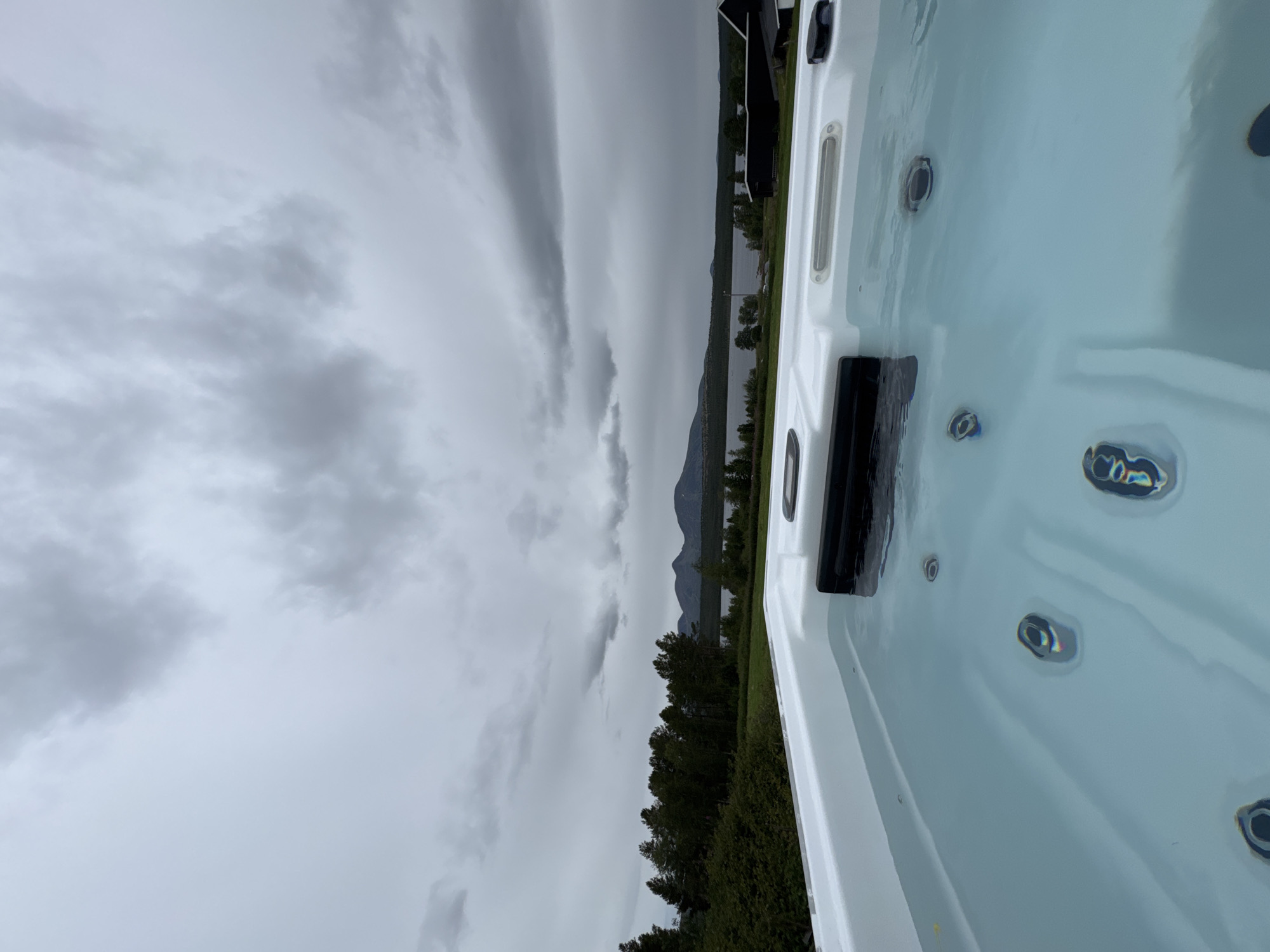 The image shows a close-up of the edge of a white hot tub filled with clear water, with several jets visible below the surface. In the background, a serene landscape unfolds under a cloudy sky. A dense line of dark green trees stretches across the middle ground, leading to a calm body of water, likely a lake or fjord. In the distance, the silhouettes of mountains rise against the horizon. The overall scene suggests a tranquil, possibly remote, outdoor setting. The location is identified as Ulvågrenda, Norway.