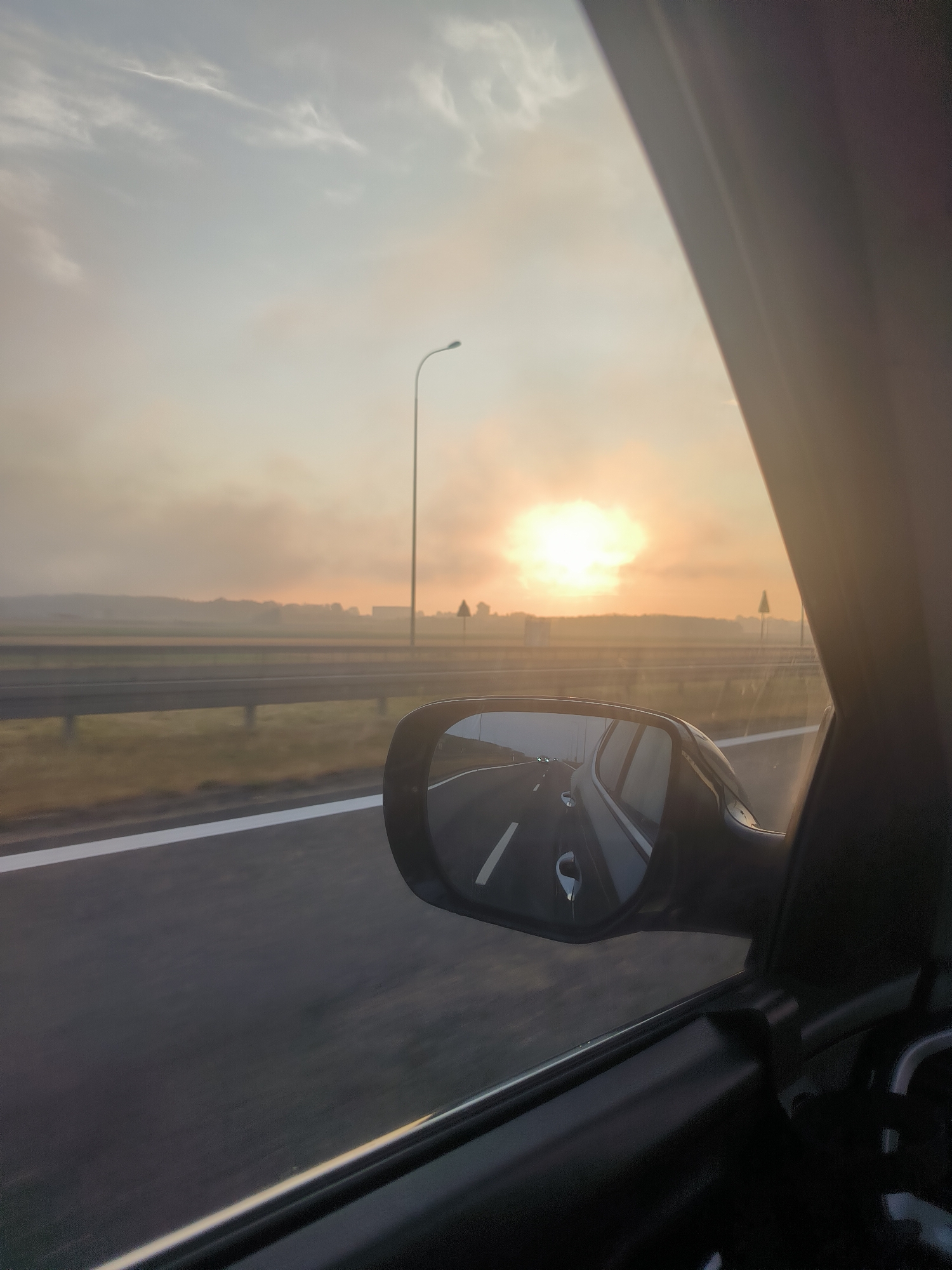 The image captures a view from inside a vehicle traveling on a highway near Olendry, Poland, during sunrise. The sun, a bright, luminous orb, is low on the horizon, casting a warm, orange glow across the sky and through a thin layer of haze or fog that obscures the distant landscape. The highway stretches ahead, with lanes marked by white lines, and a guardrail runs alongside. A street lamp is visible on the left side of the road. In the side mirror of the car, the receding highway and other vehicles are visible, reflecting the sunlight. The interior of the car, including the window frame and part of the dashboard or door panel, is also visible in the foreground, framing the outdoor scene.
