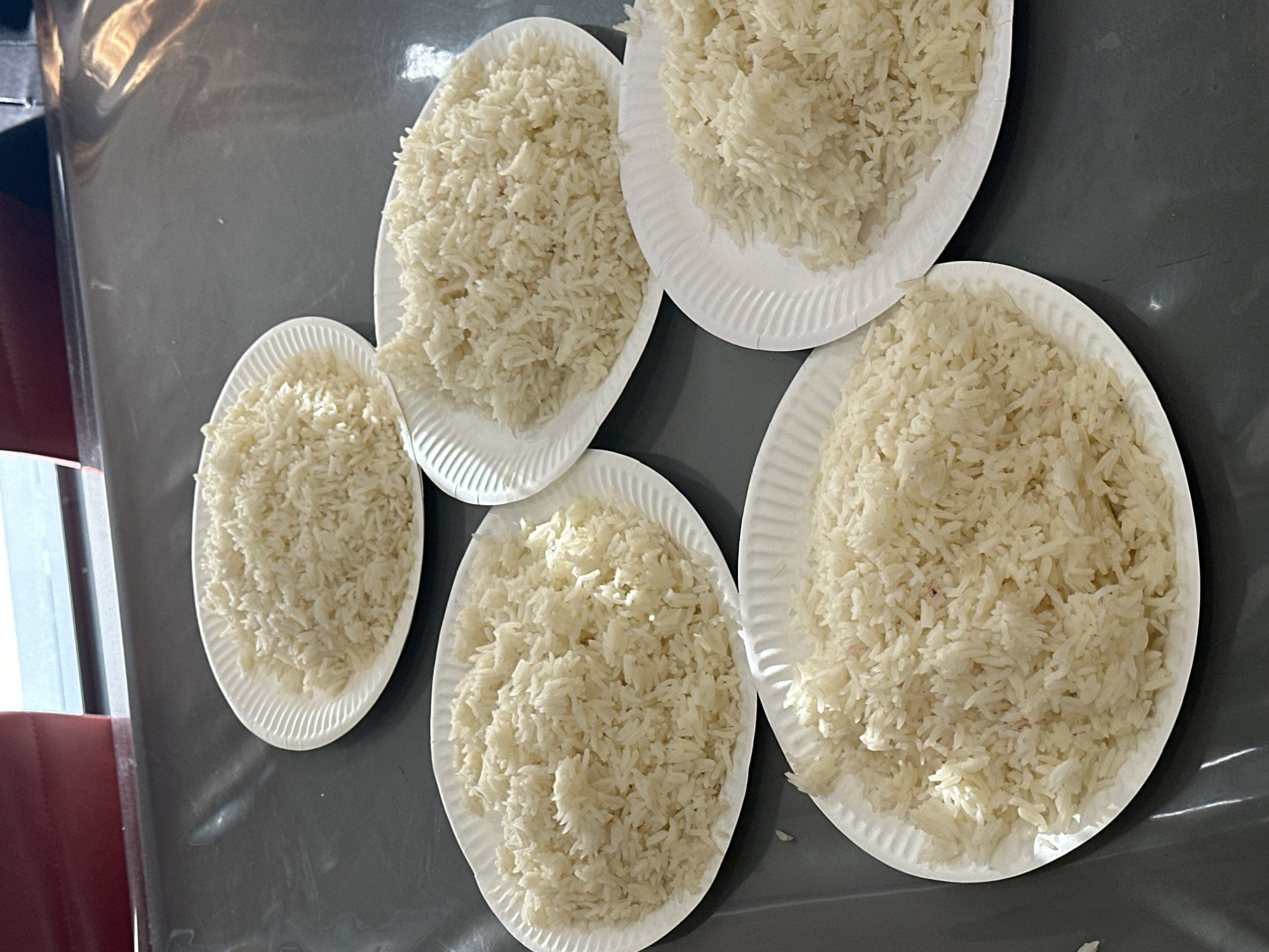 The image depicts five plates of white rice, each on a white paper plate with a ridged edge. The plates are arranged on a dark gray, possibly glossy, surface, likely a table or countertop. A red chair or object is partially visible in the upper left corner of the image. The location of the image is Yerevan, Armenia.