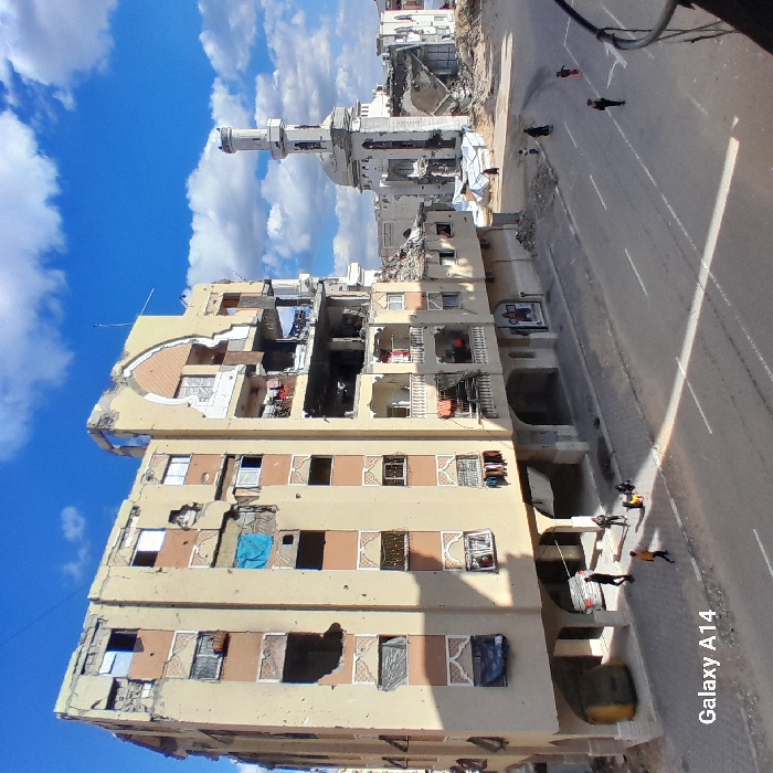 The image displays an urban street scene in Khan Yunis, Palestinian Territory, characterized by significant structural damage under a clear blue sky with scattered white clouds. A prominent multi-story (approximately 6-floor) building, light beige with peach-colored accent panels, dominates the left-midground. This building exhibits extensive damage, including large impact craters, exposed interiors, shattered windows, and numerous pockmarks on its facade. Clothing hangs from several balconies and exposed window openings, with a blue tarp visible from an upper floor. The ground level features an open arcade. To the right, in the background, a white mosque with a dome and a tall, cylindrical minaret is heavily damaged, with large sections of its walls and roof collapsed. Debris is visible around its base, and a white tarp covers an area nearby. A paved road with white lane markings runs across the mid-ground. On the left side of the street, under the damaged building, a yellow utility vehicle is visible, along with approximately four individuals on foot. On the right side of the street, an additional four figures, including adults and children, are seen walking along the roadway. The scene is brightly lit by sunlight, casting visible shadows.